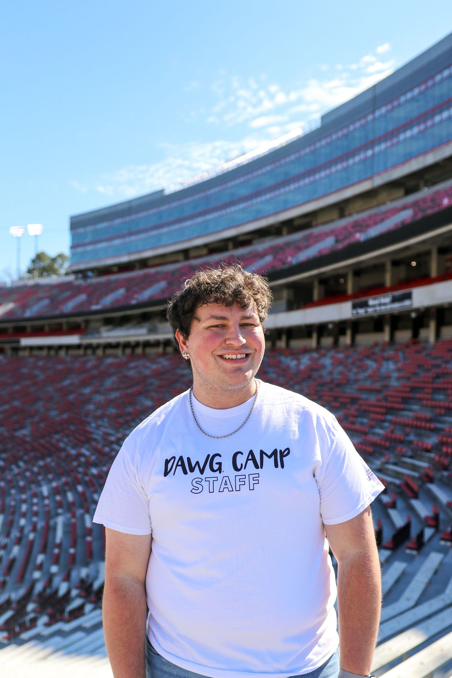 A person wearing a white 'DAWG CAMP STAFF' t-shirt stands in an empty stadium with red seats on a sunny day.