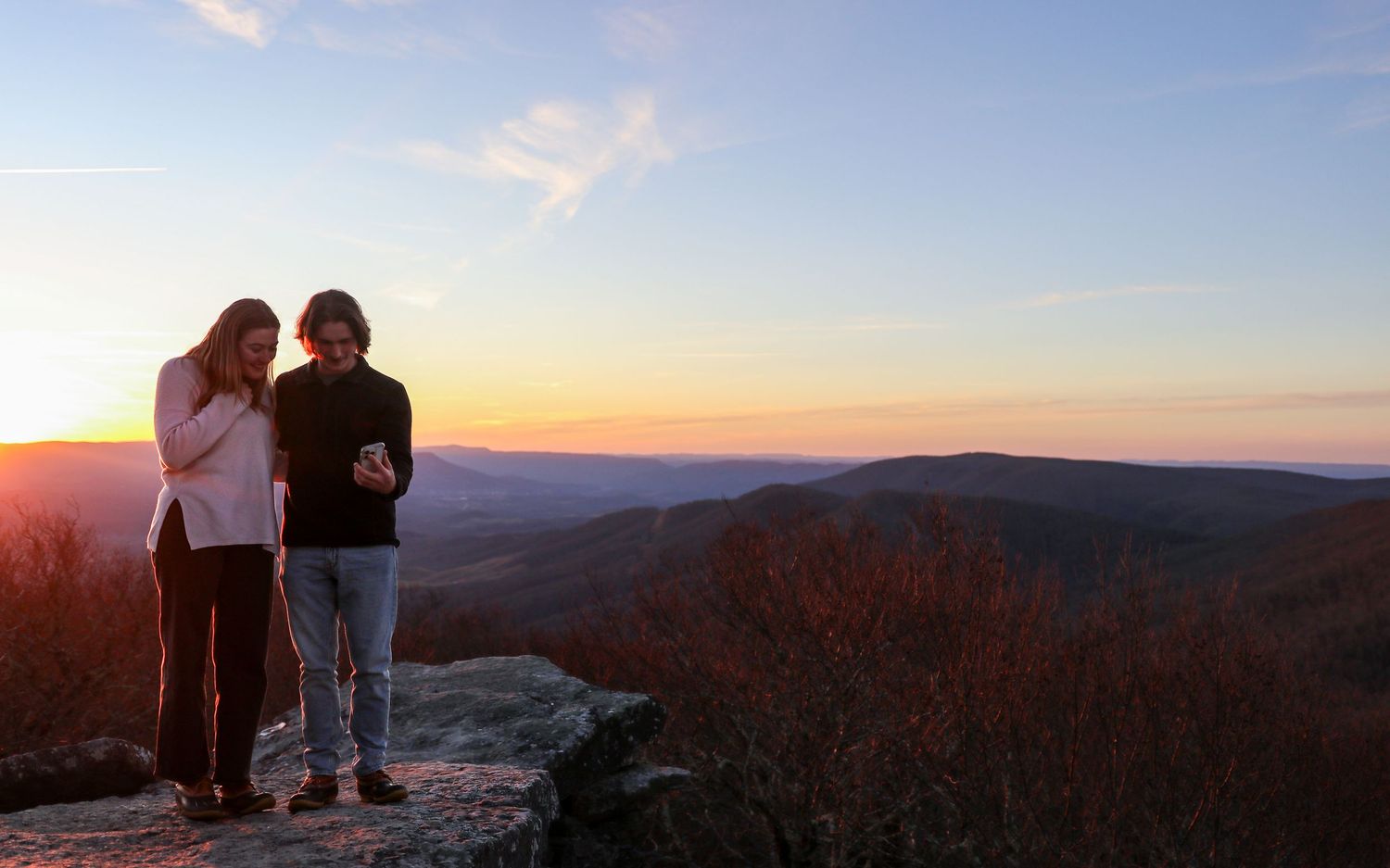 Two people stand on a mountain overlook during a colorful sunset over rolling hills and distant peaks.