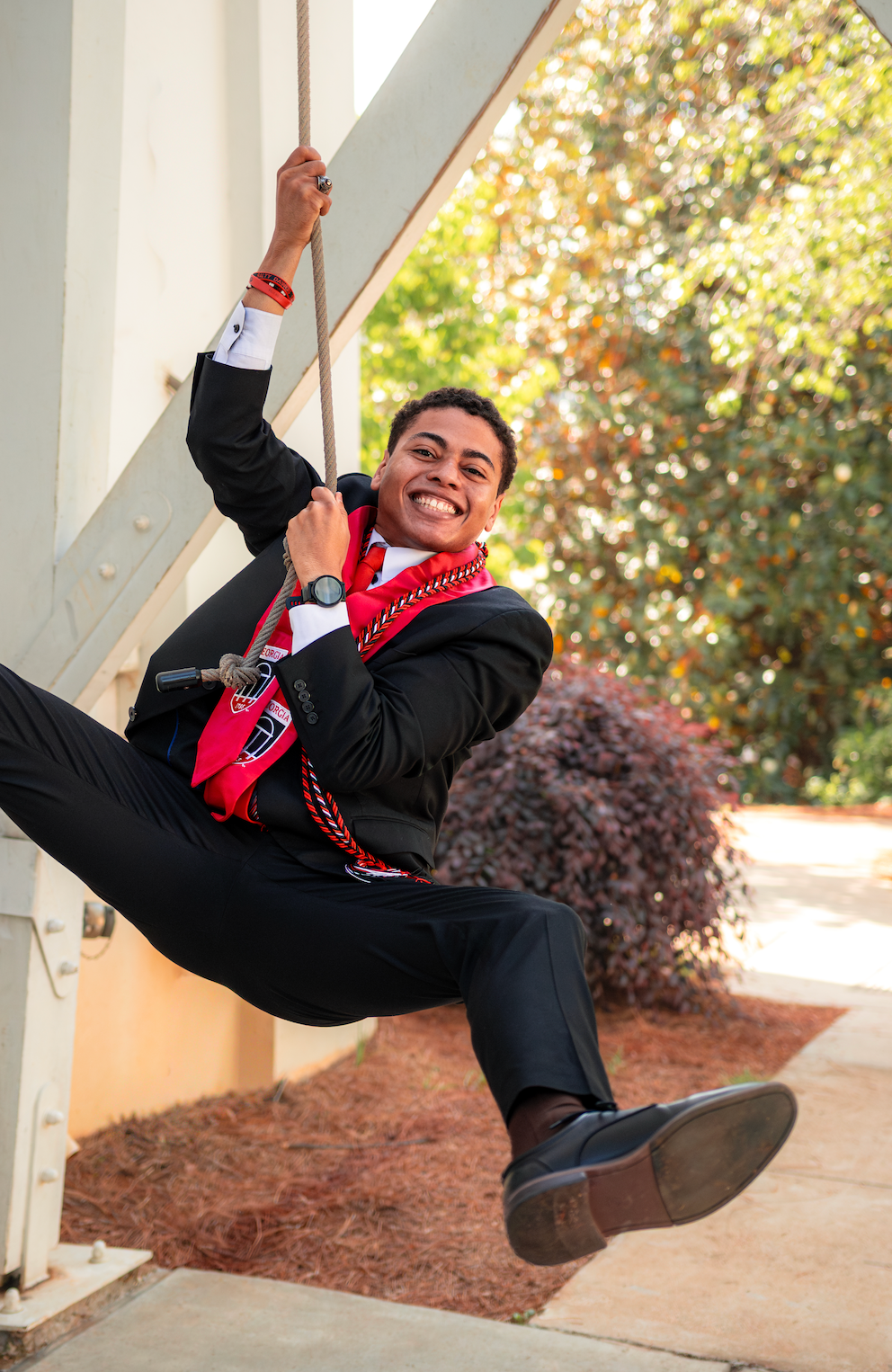 Smiling young man in formal attire with red sash hangs from a rope outdoors, posing playfully near a building.