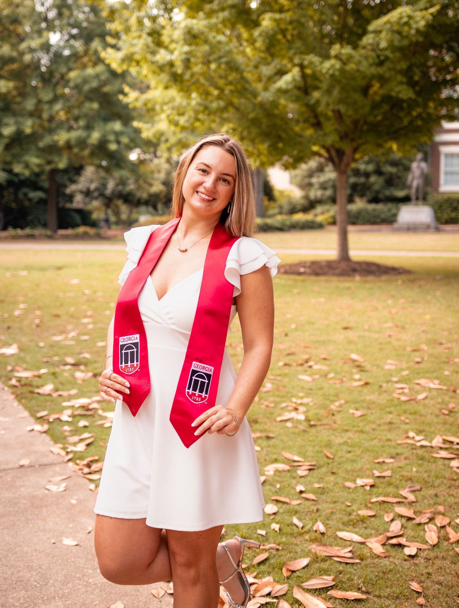 A smiling young woman in a white dress wearing a red graduation stole poses outdoors on a college campus.