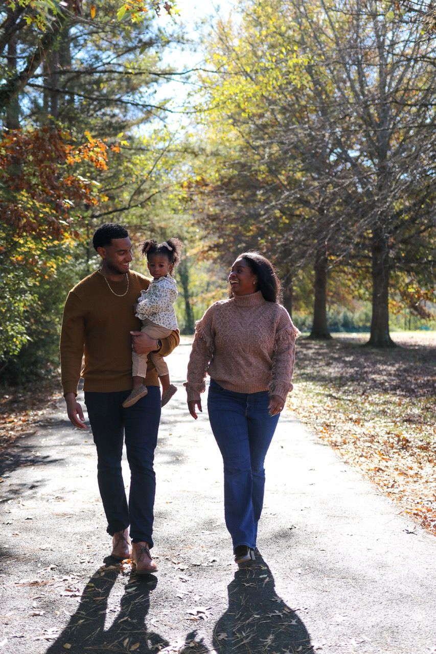 A couple walks together on a sunlit autumn path surrounded by trees with changing leaves and falling foliage.