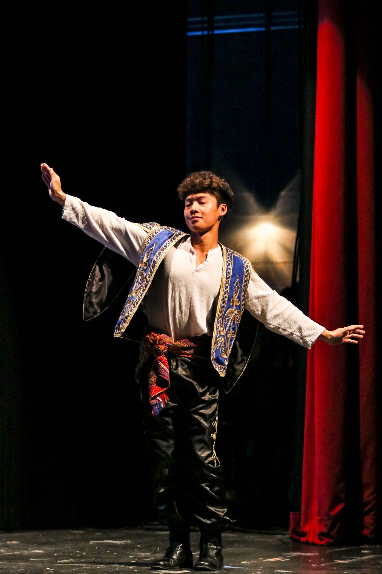 A performer in traditional folk costume dances expressively on a dark stage with red curtains in the background.