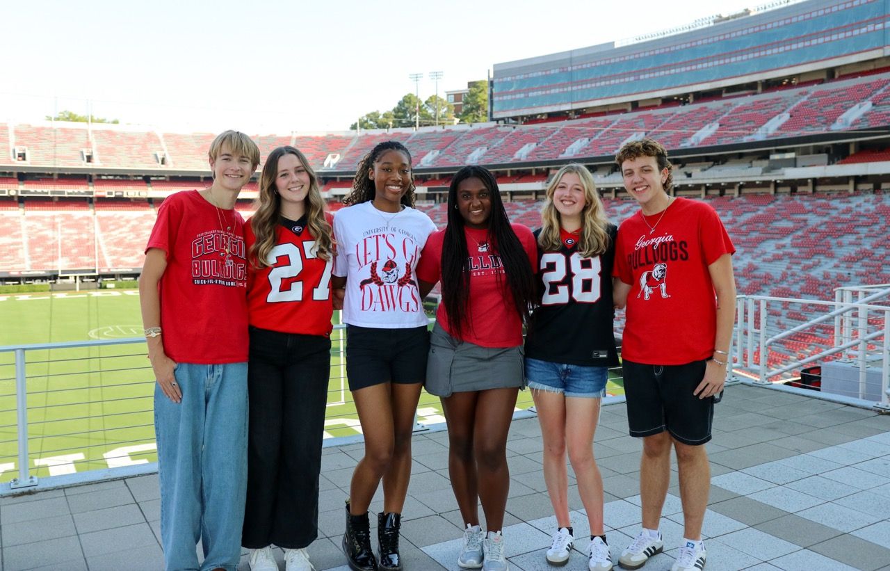 A group of students stand together in athletic wear on the bleachers of a large football stadium.