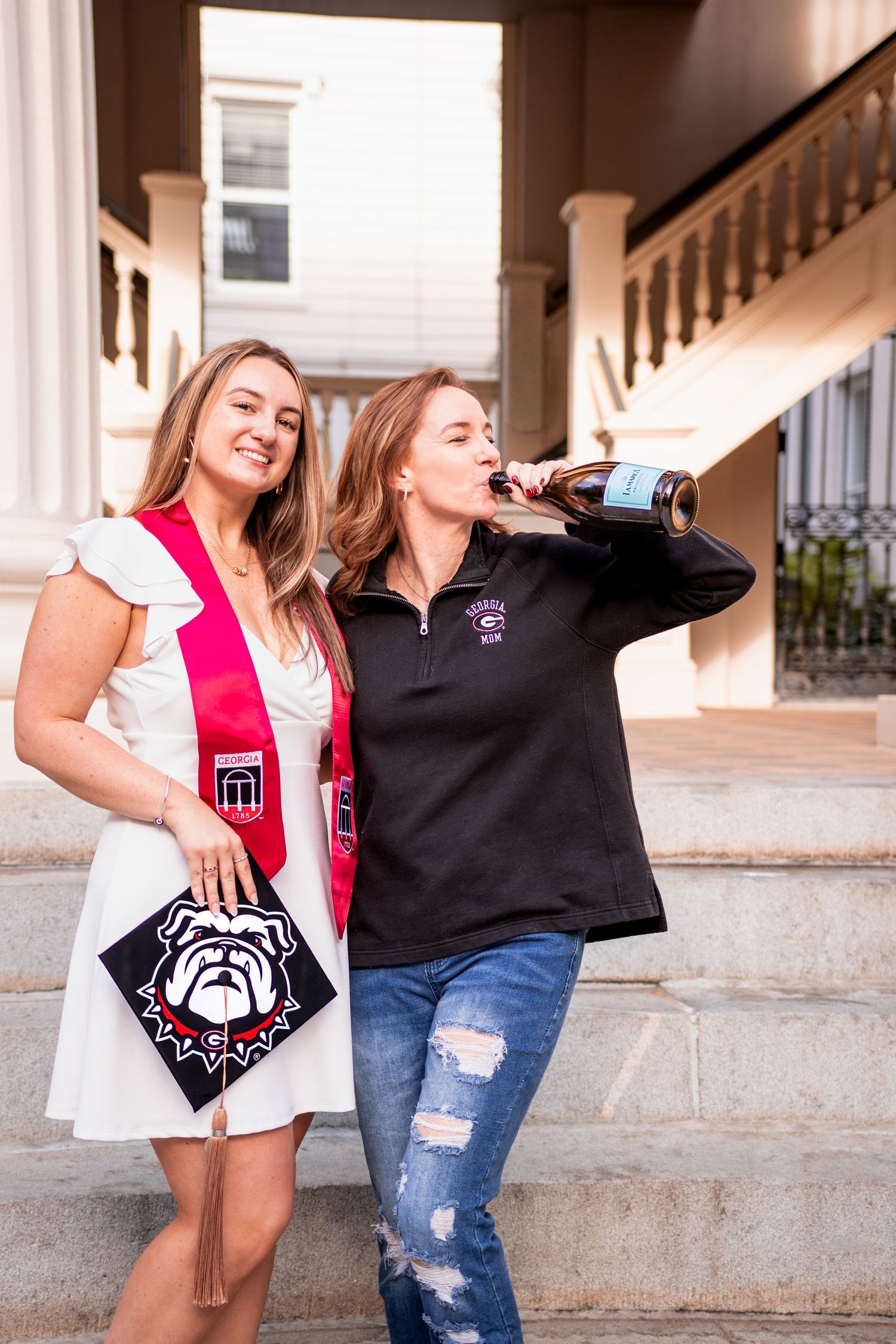 Two college graduates celebrate outdoors on steps, one wearing a red sash and holding a bulldog graduation cap.