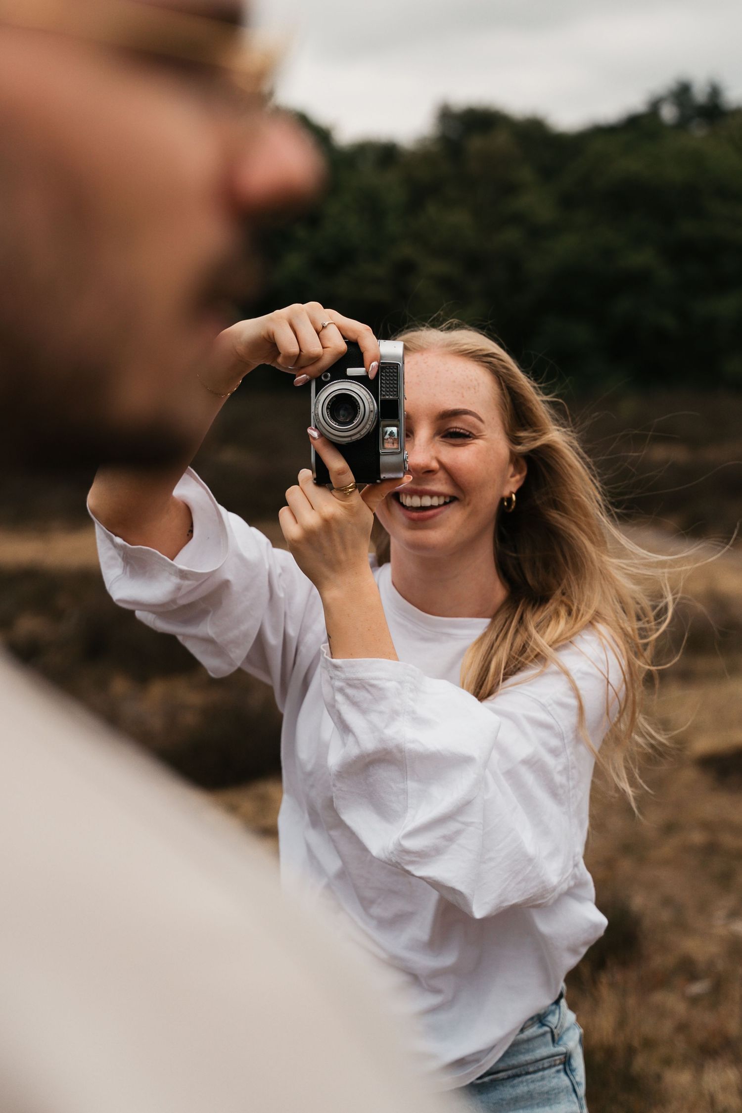 Een verliefd stel op de paarse heide in Laren tijdens een frisse lentedag, terwijl een persoon met een camera vrolijk poseert.