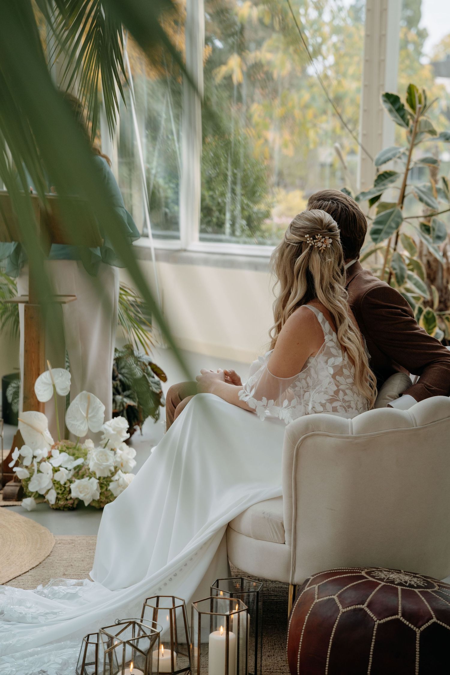 A bride in a flowing white lace gown sits gracefully on an elegant cream-colored couch in a sunlit room.