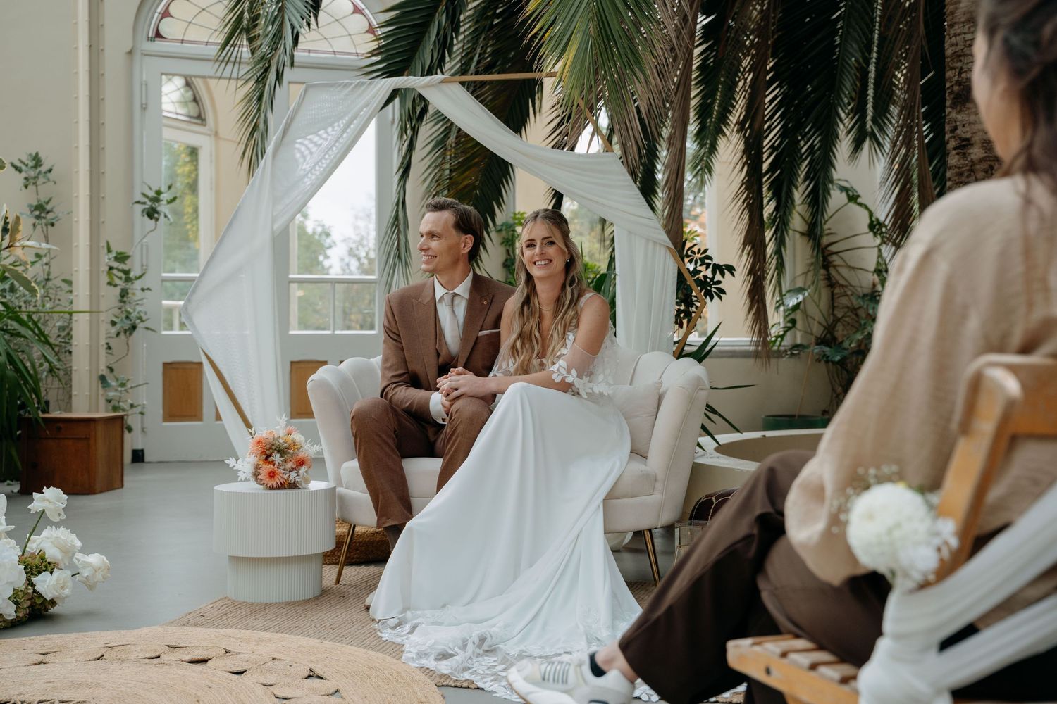 A couple sits together in a bright indoor garden space during an intimate wedding ceremony.