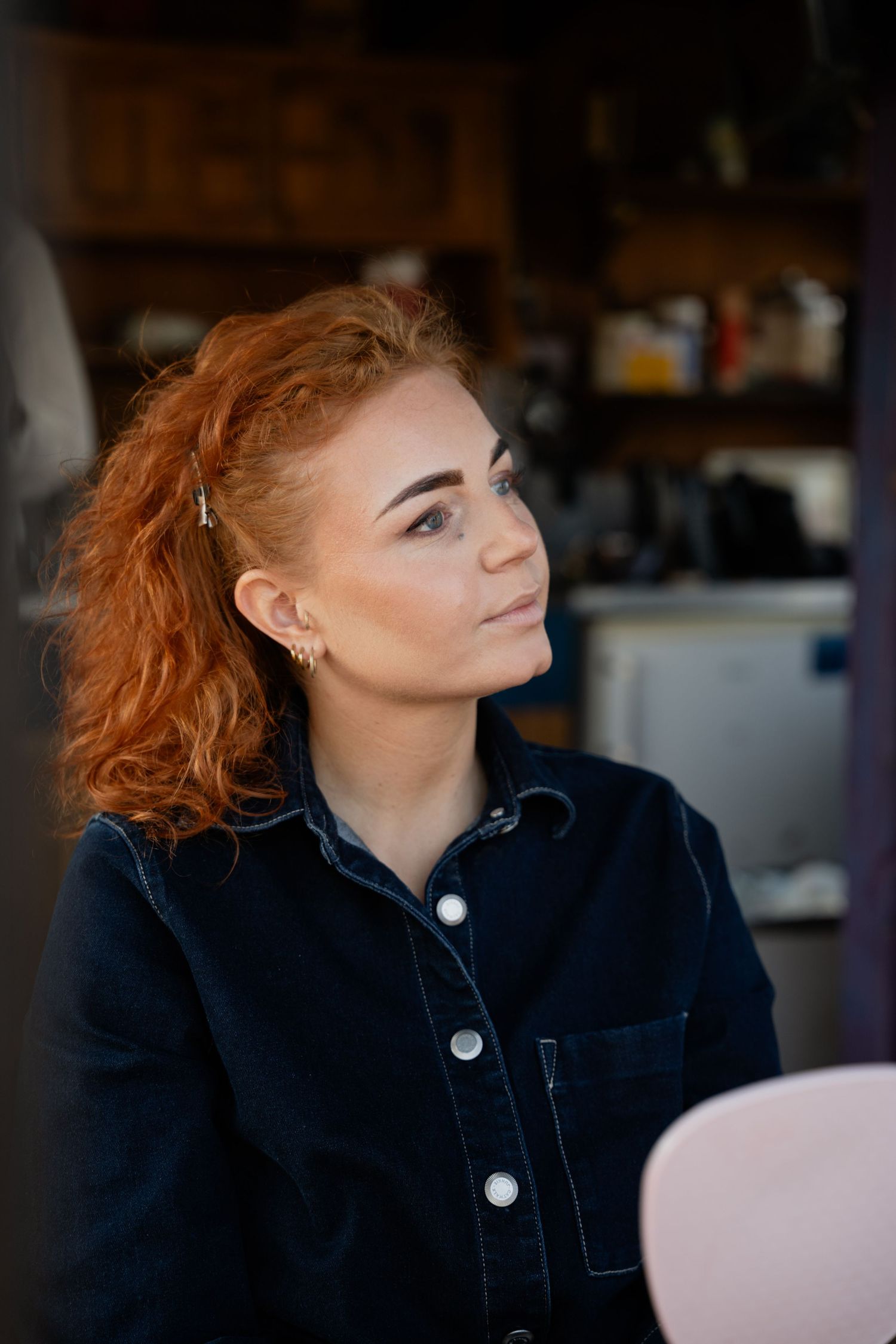 Young woman with curly red hair wearing a dark denim jacket, sitting indoors with a thoughtful expression.