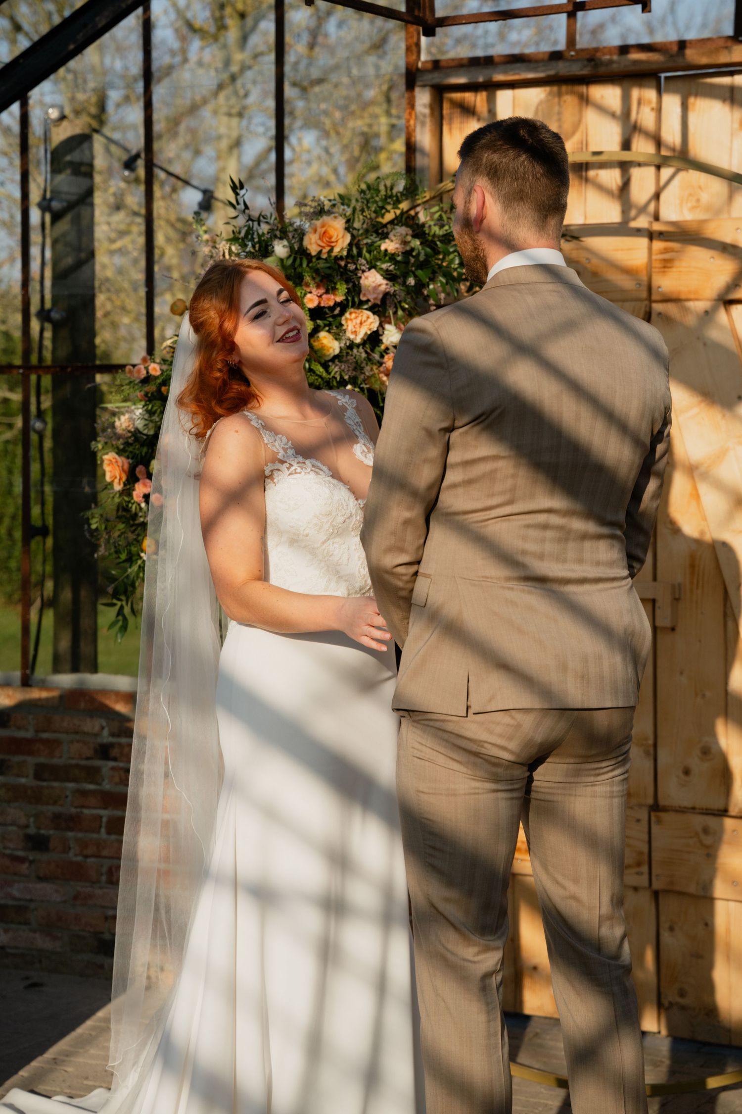 Bride in white lace gown and veil smiles at groom in tan suit during outdoor wedding ceremony with floral arch.