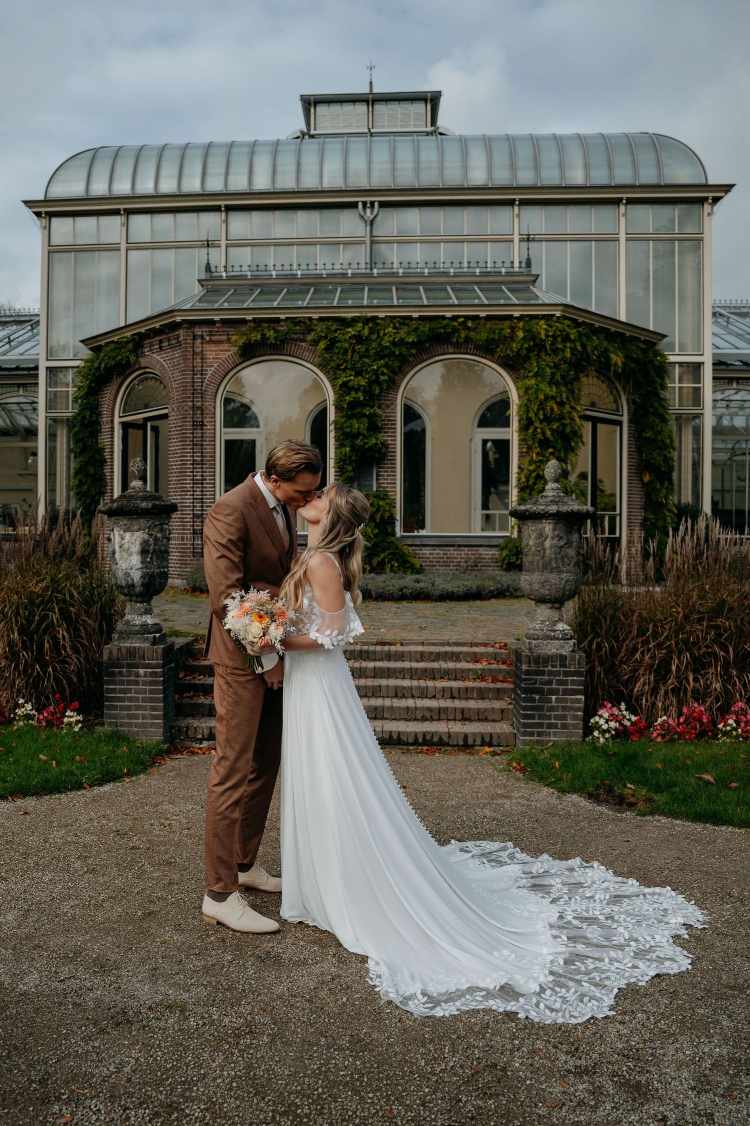 A romantic wedding couple embraces in front of an elegant greenhouse conservatory with ornate architecture.