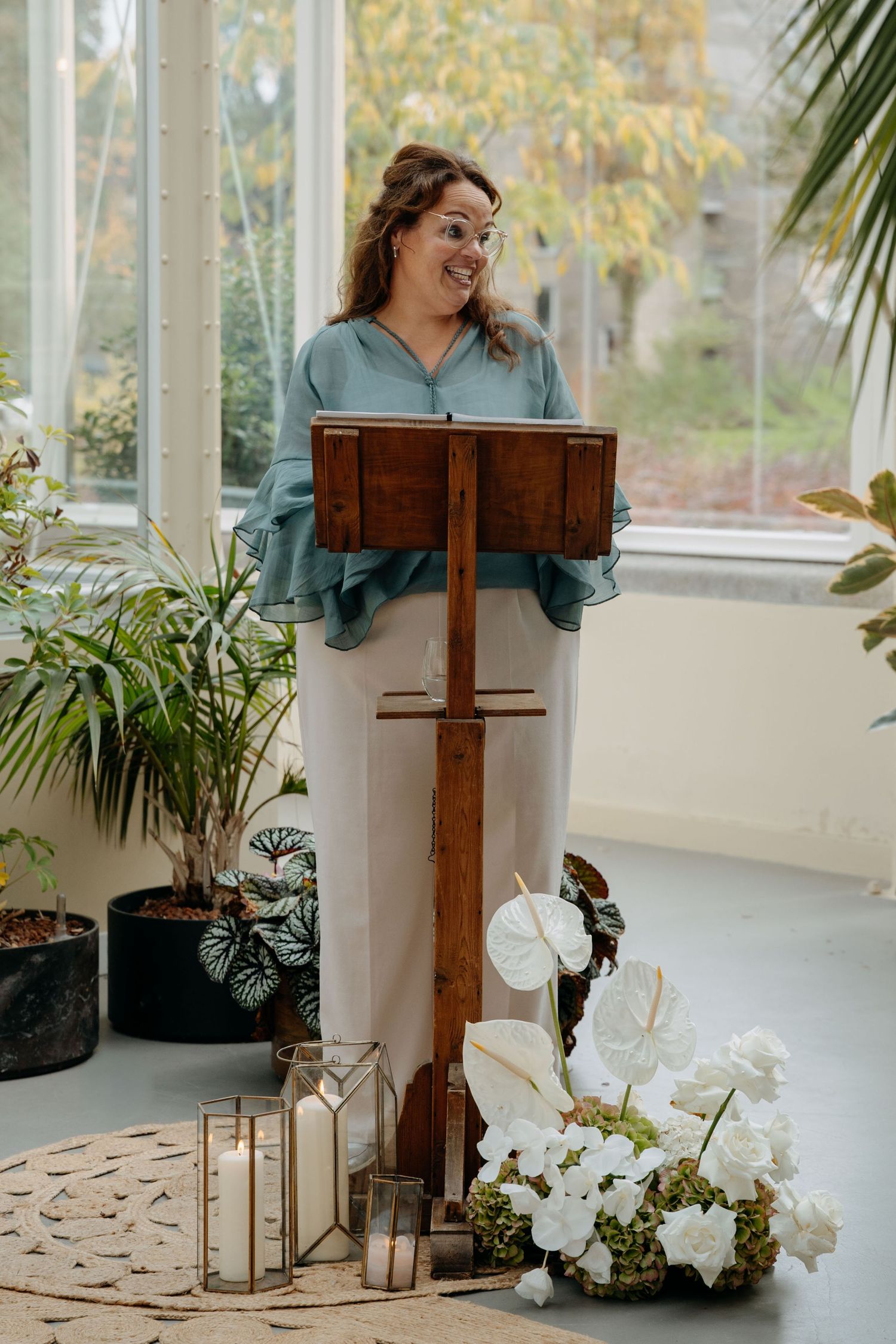 A speaker stands at a wooden podium surrounded by white orchids and lantern decorations in a sunlit conservatory setting.