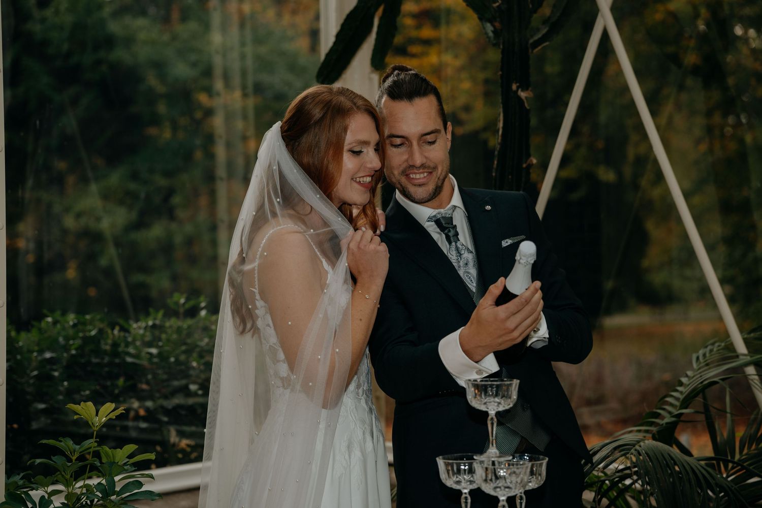 Bride and groom smiling together during outdoor wedding ceremony, bride wearing veil, groom in navy suit holding champagne bottle.