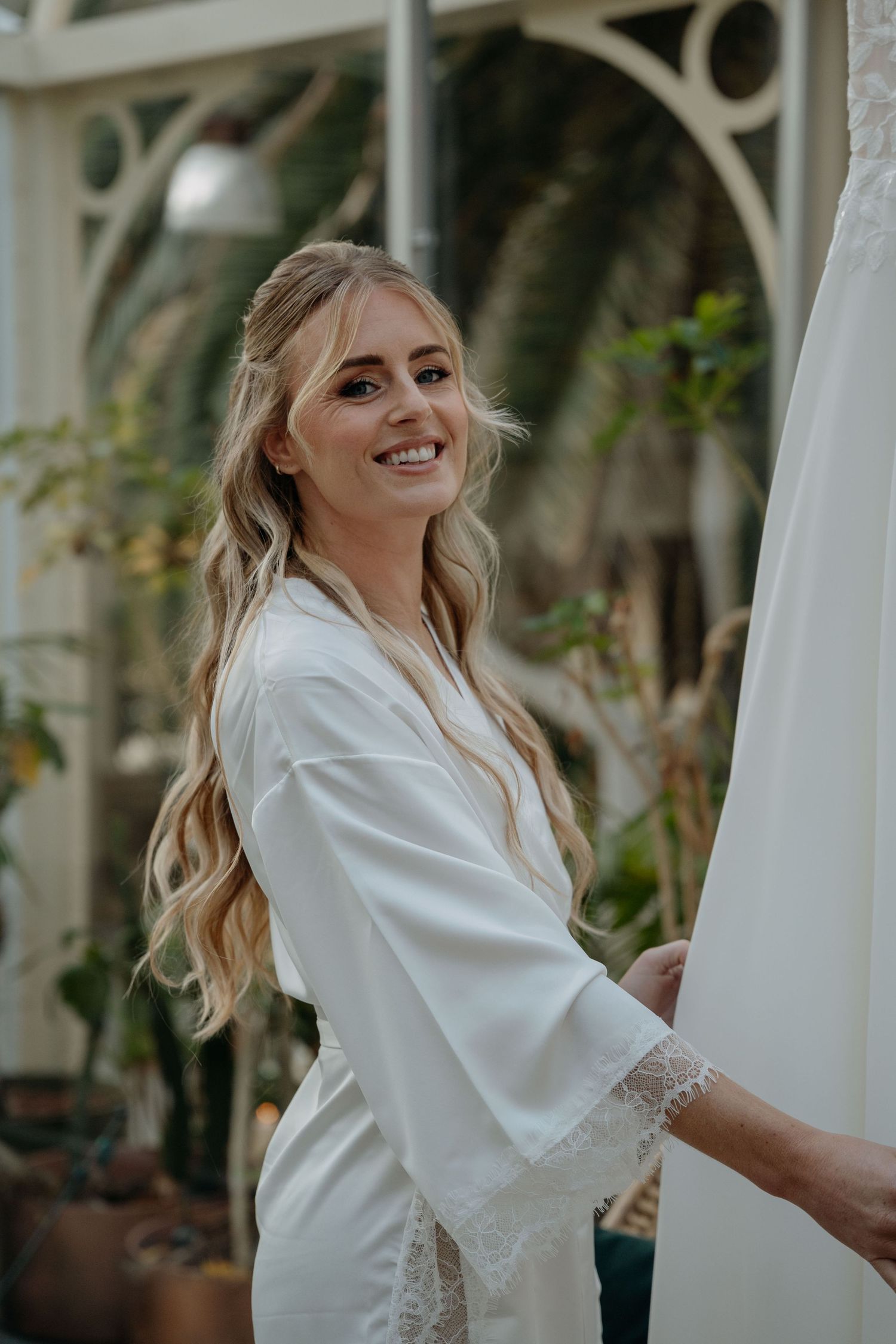 Bride in white silk robe smiles joyfully while getting ready on her wedding morning in a bright sunlit room.