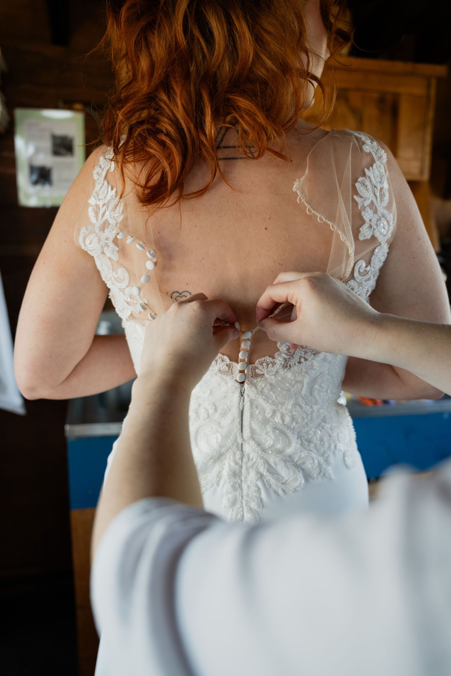 Helper fastening buttons on back of lace wedding dress worn by red-haired bride during bridal preparations.