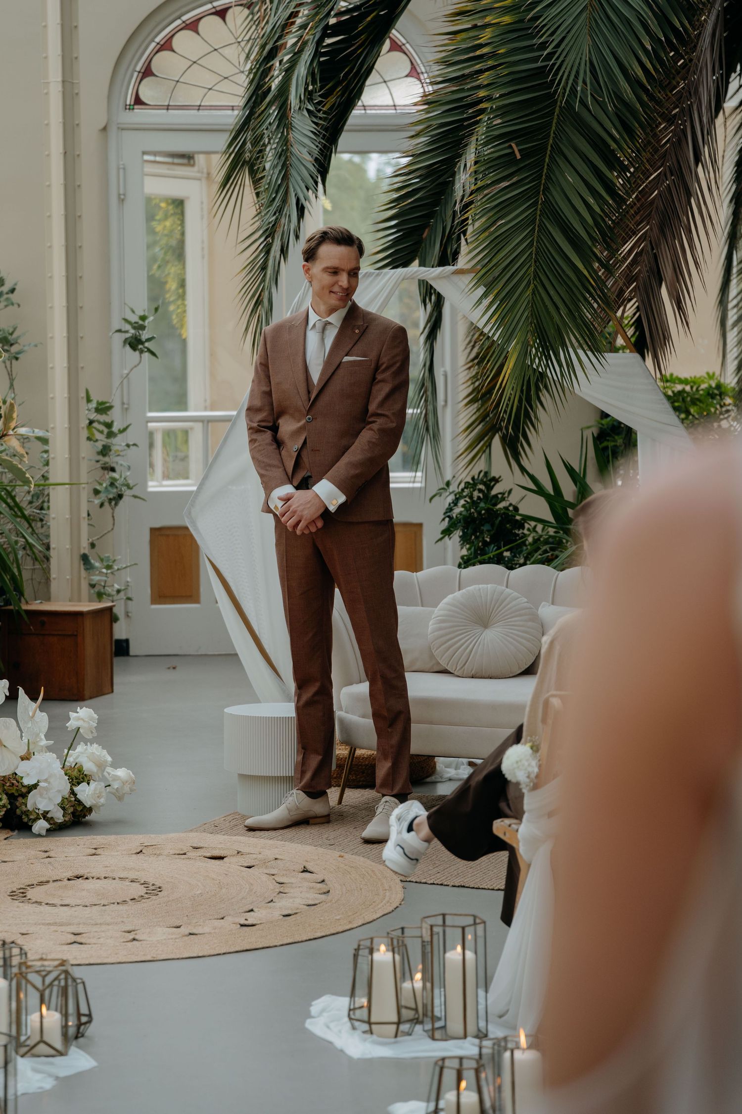 A brown-suited figure stands in an elegant indoor space with palm fronds and white decor during a wedding ceremony.