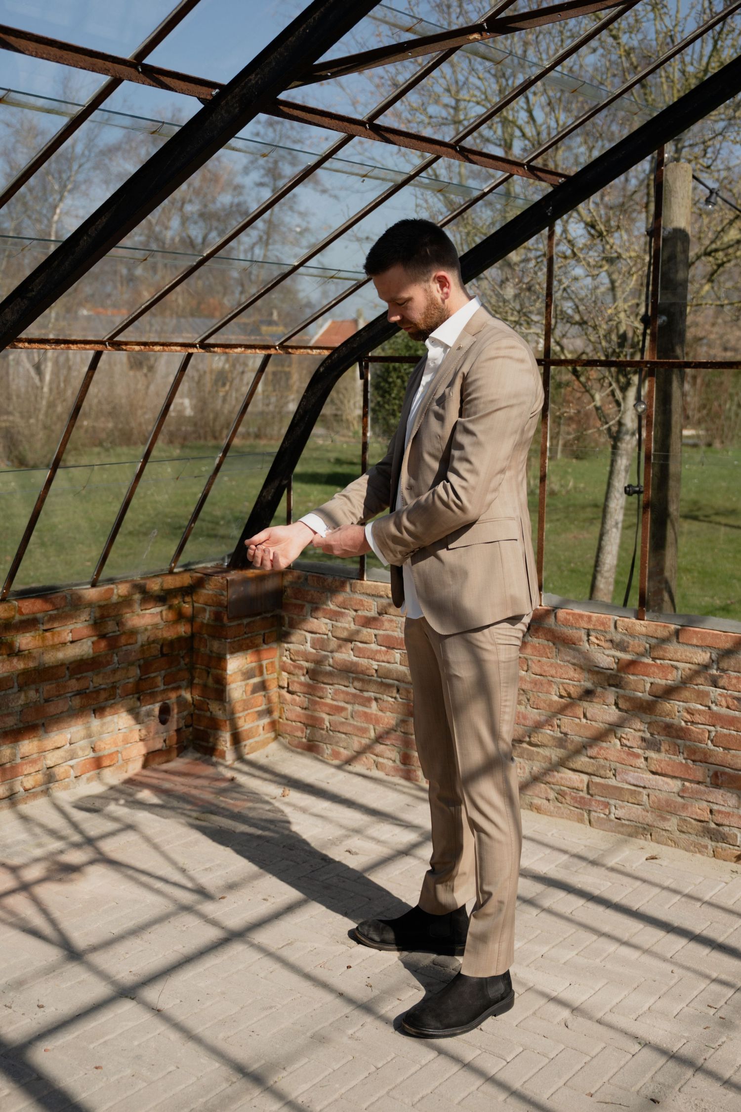 Man in beige suit adjusting cufflinks inside rustic greenhouse with brick walls and metal frame structure.