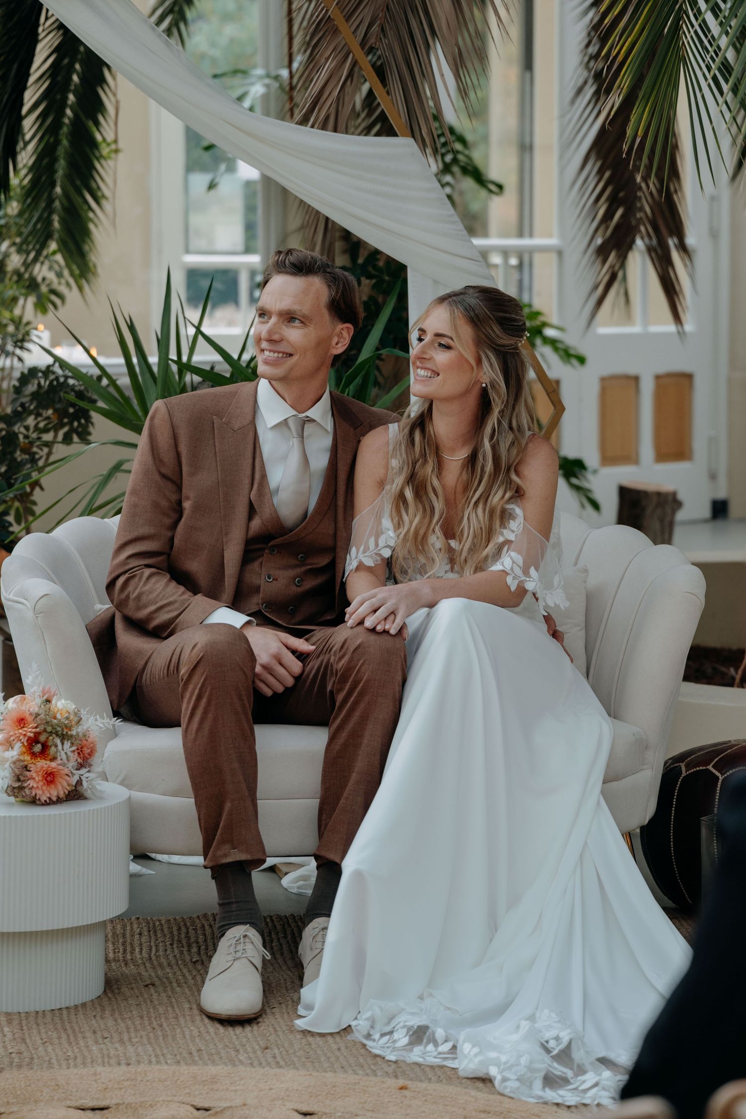 A couple shares a joyful moment on a white couch surrounded by tropical plants in a bright indoor setting.