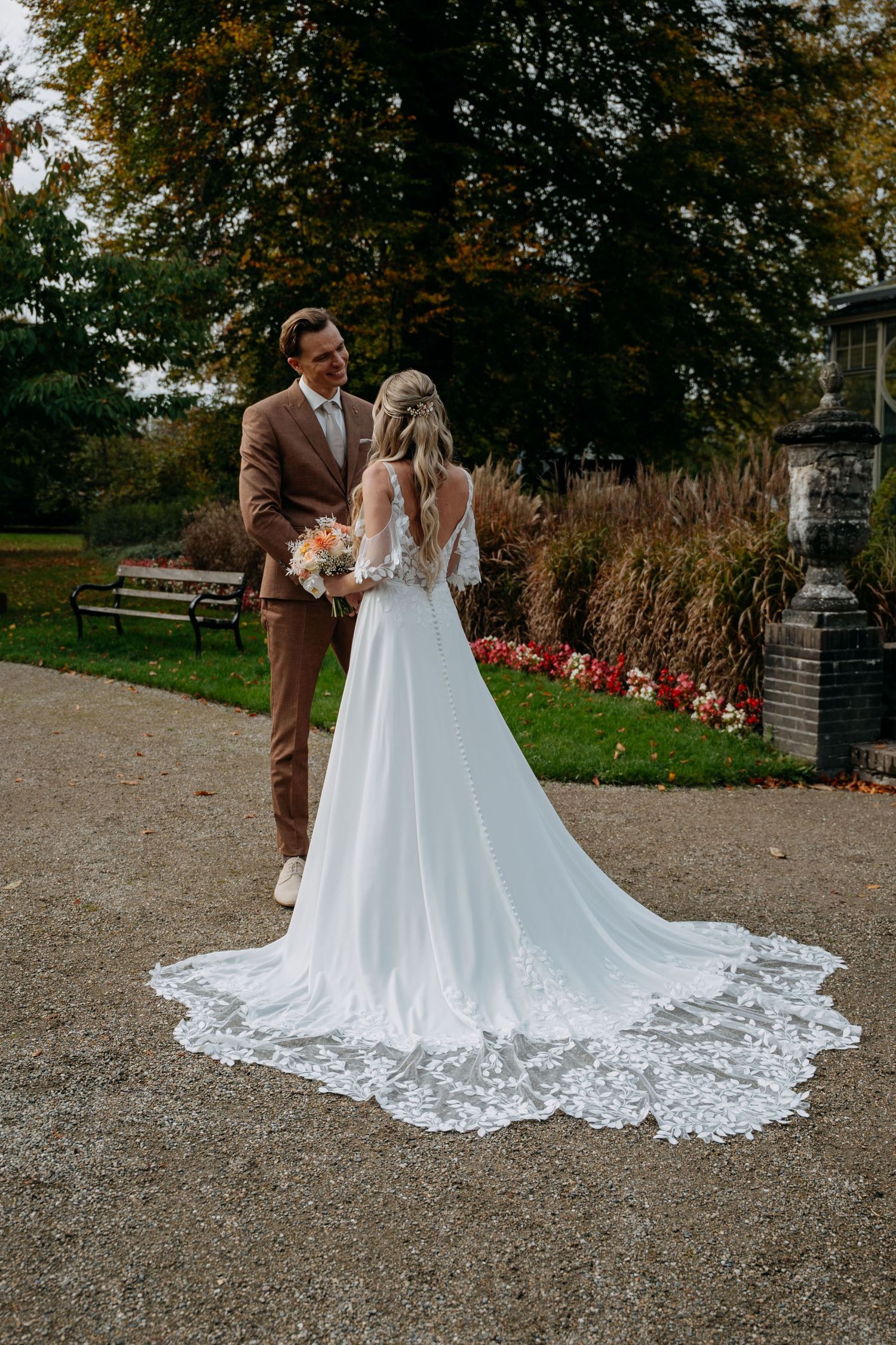 A couple in wedding attire embraces outdoors, with the bride wearing a flowing white gown with intricate lace train details.