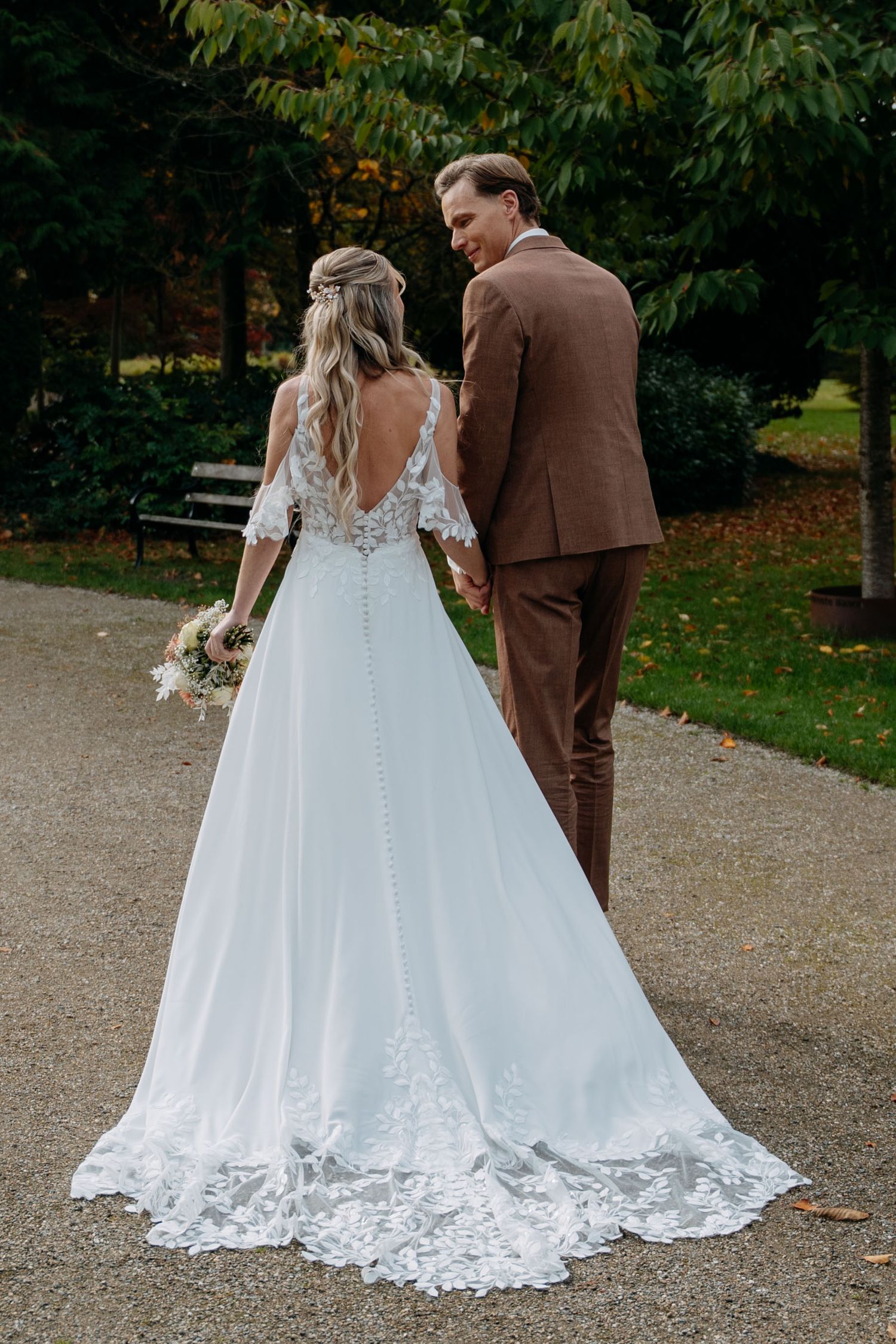 A bride in a lace-detailed white gown stands with her partner in a brown suit on a garden path during autumn.