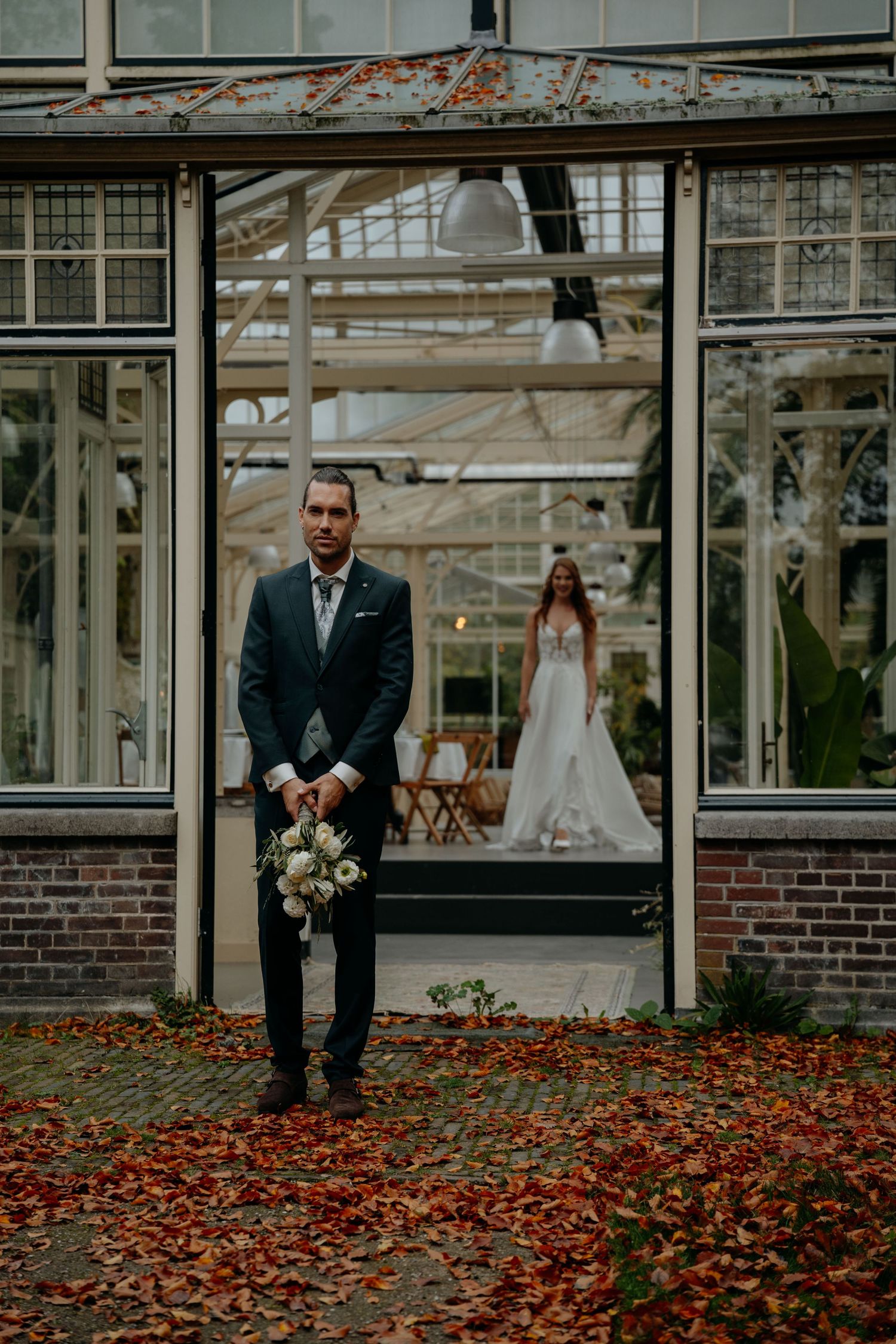 Groom holding bouquet outside greenhouse with bride visible through glass doors, autumn leaves on ground.