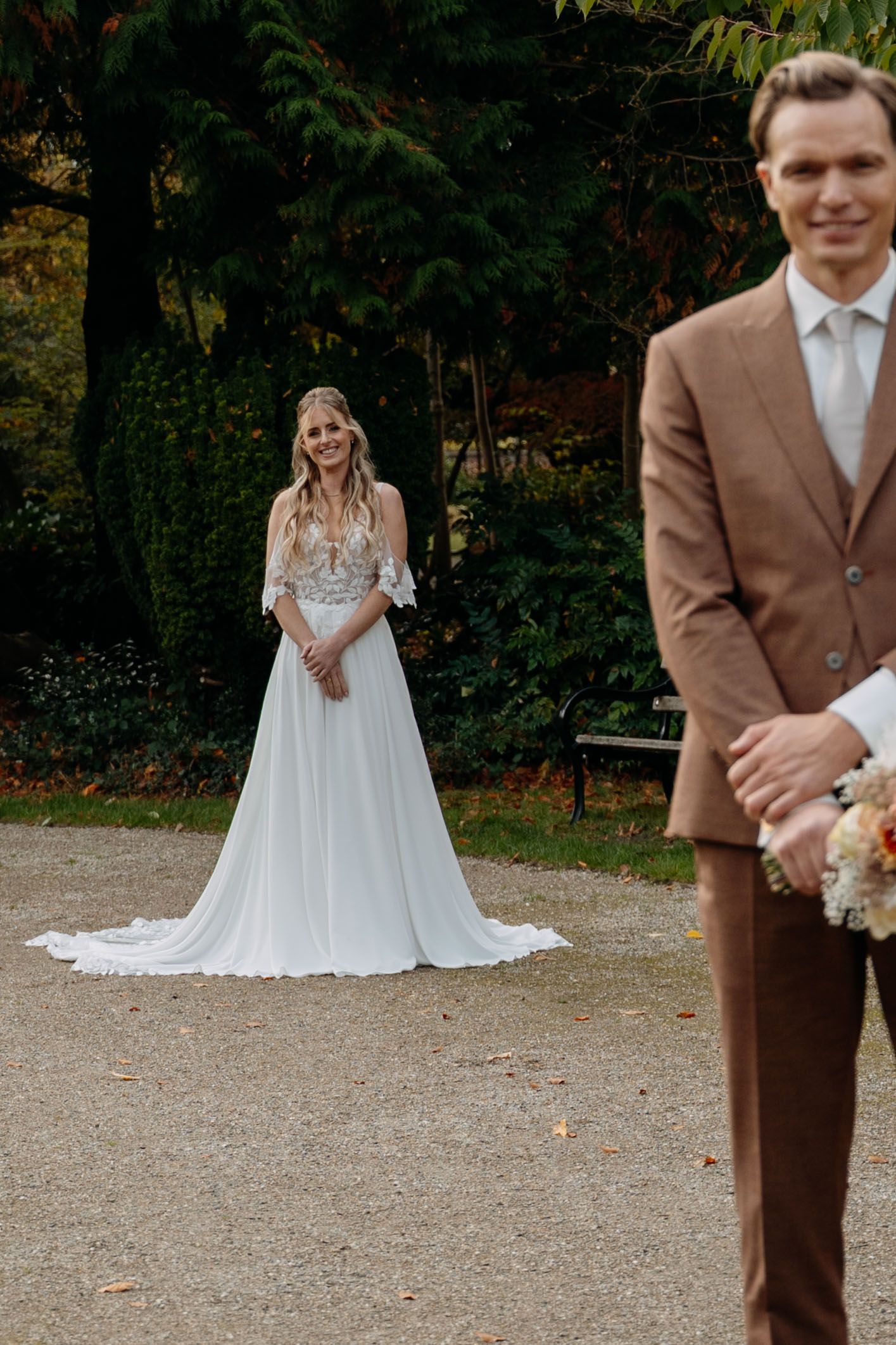 Wedding couple poses outdoors with bride in flowing white gown and groom in brown suit on gravel path near trees.