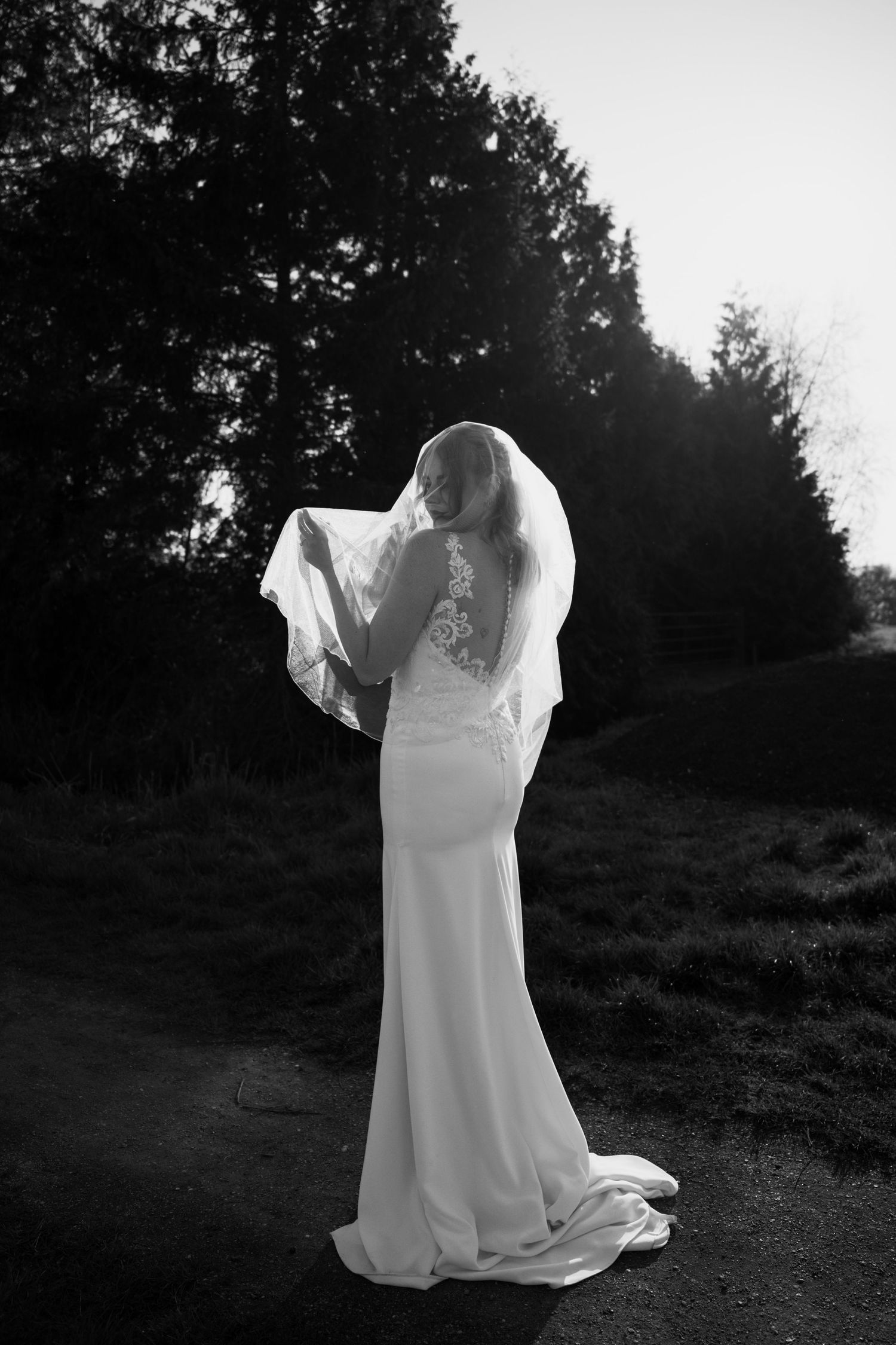 Black and white photo of a bride in a white gown and veil standing outdoors among trees.