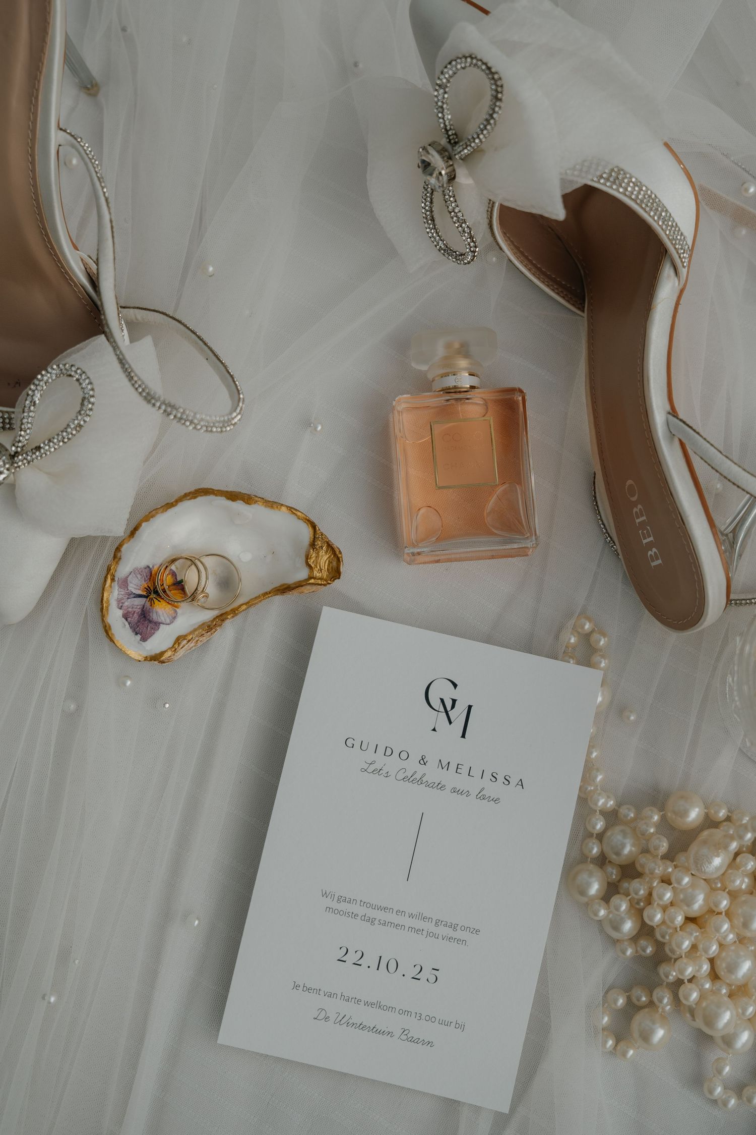Wedding accessories including pearls, perfume, and invitation cards arranged on white fabric.
