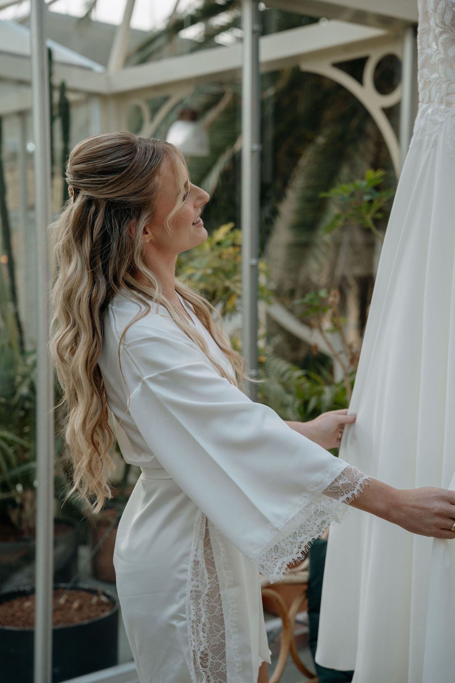 Woman in white lace robe touches wedding dress in greenhouse setting with natural light streaming through windows.