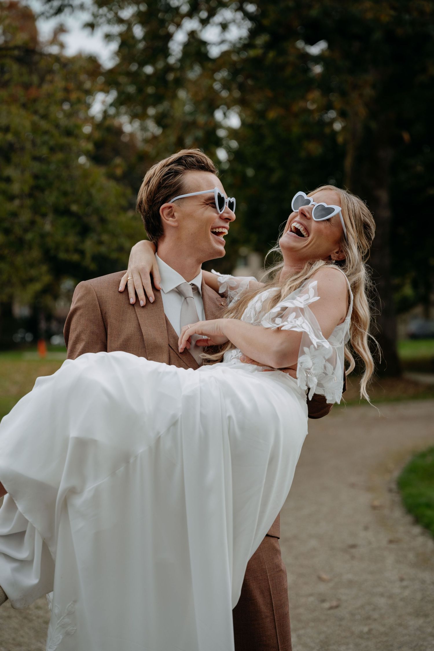 A joyful couple in wedding attire laughing together while posing playfully on a tree-lined path during autumn.