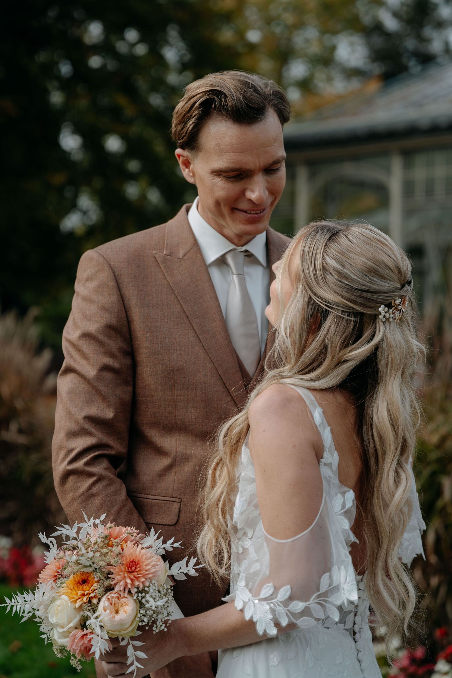 A couple shares an intimate moment in a garden setting, with the bride holding a bouquet of peach and coral flowers.