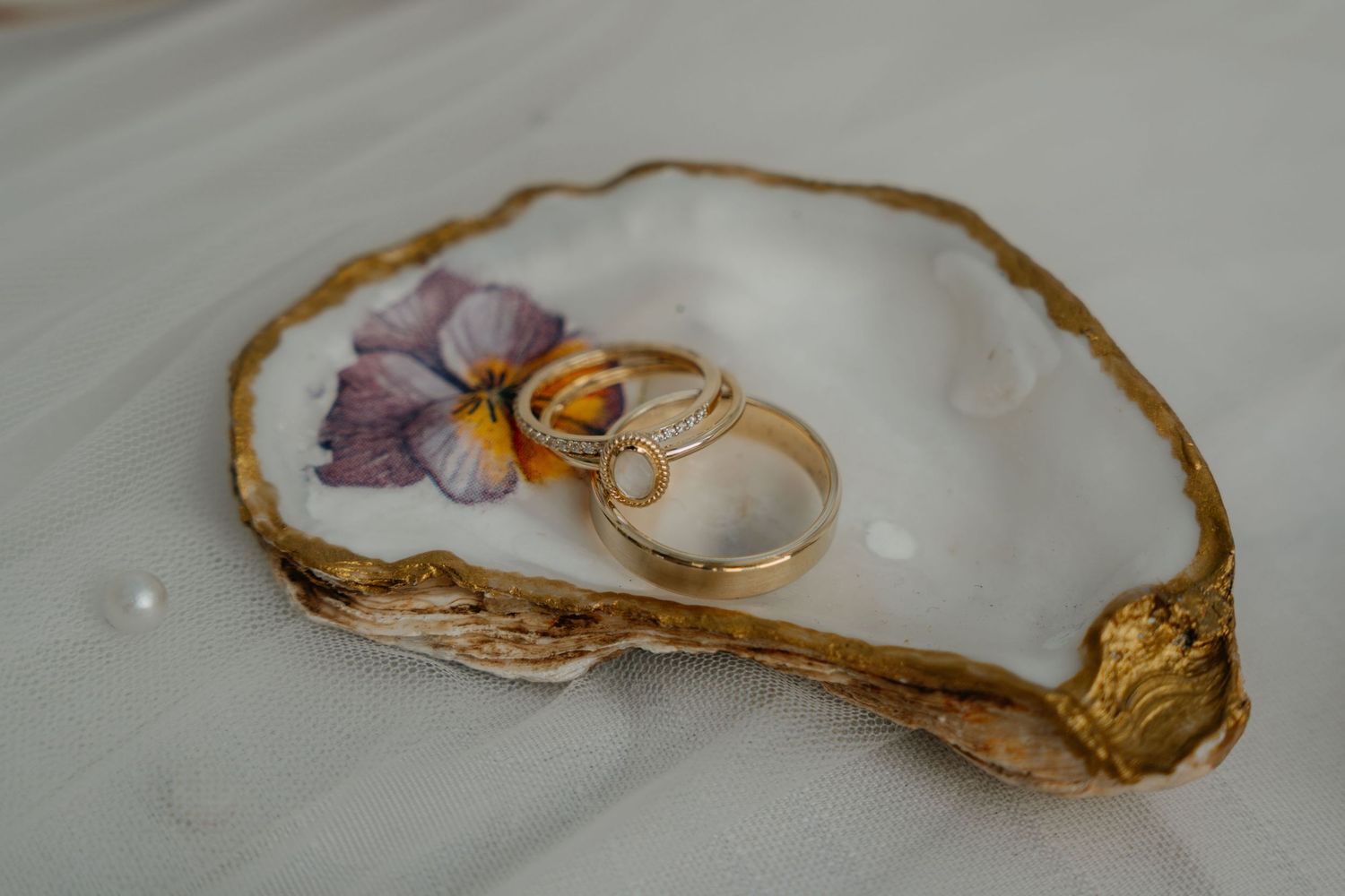 Wedding rings displayed on an oyster shell with purple flowers and gold accents on a white surface.