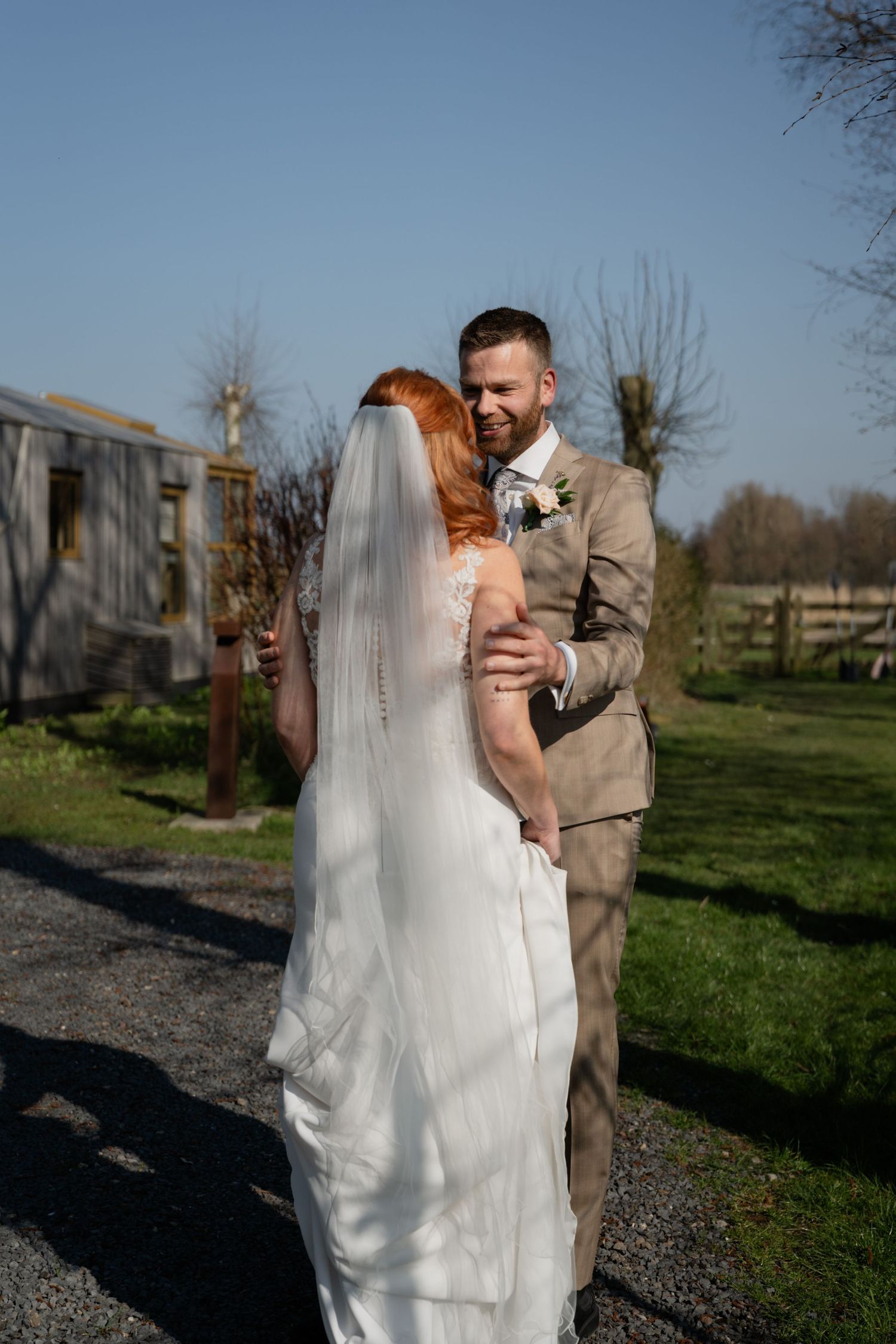 Groom in tan suit smiling at red-haired bride in white gown and veil during outdoor wedding ceremony.