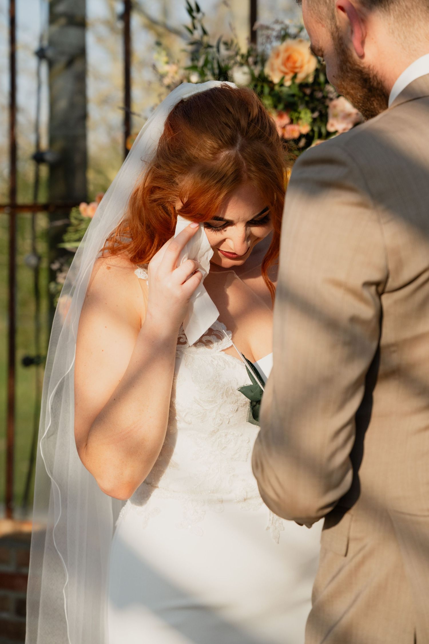 Emotional bride in white gown and veil wiping tears during outdoor wedding ceremony with groom in tan suit.
