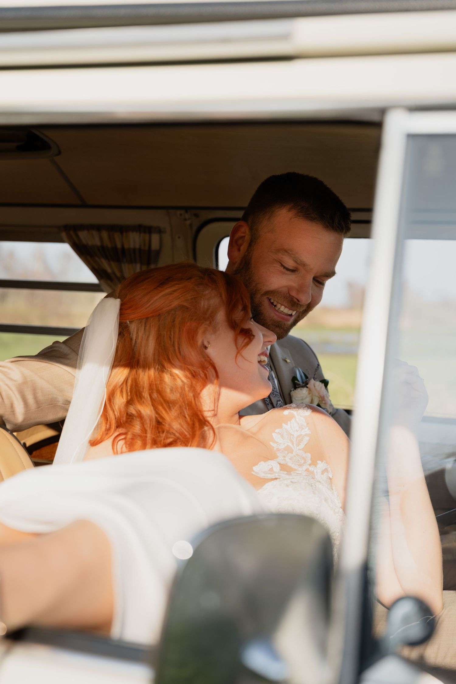 A smiling groom embraces a redhead bride in a vintage van on their wedding day.