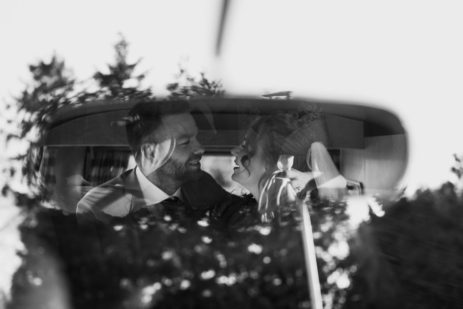 Black and white photo of a couple sharing a joyful moment inside a vintage car, reflected in the rearview mirror.