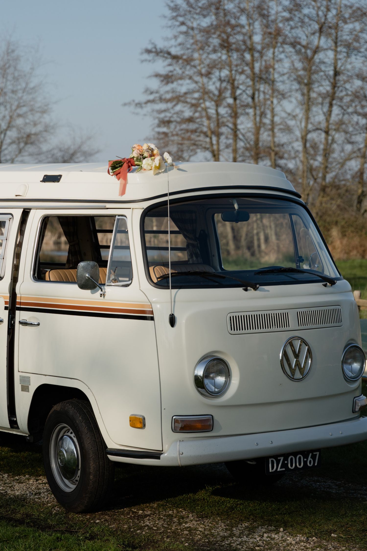 Vintage white Volkswagen Type 2 camper van with flower decoration on roof, parked outdoors with trees in background.