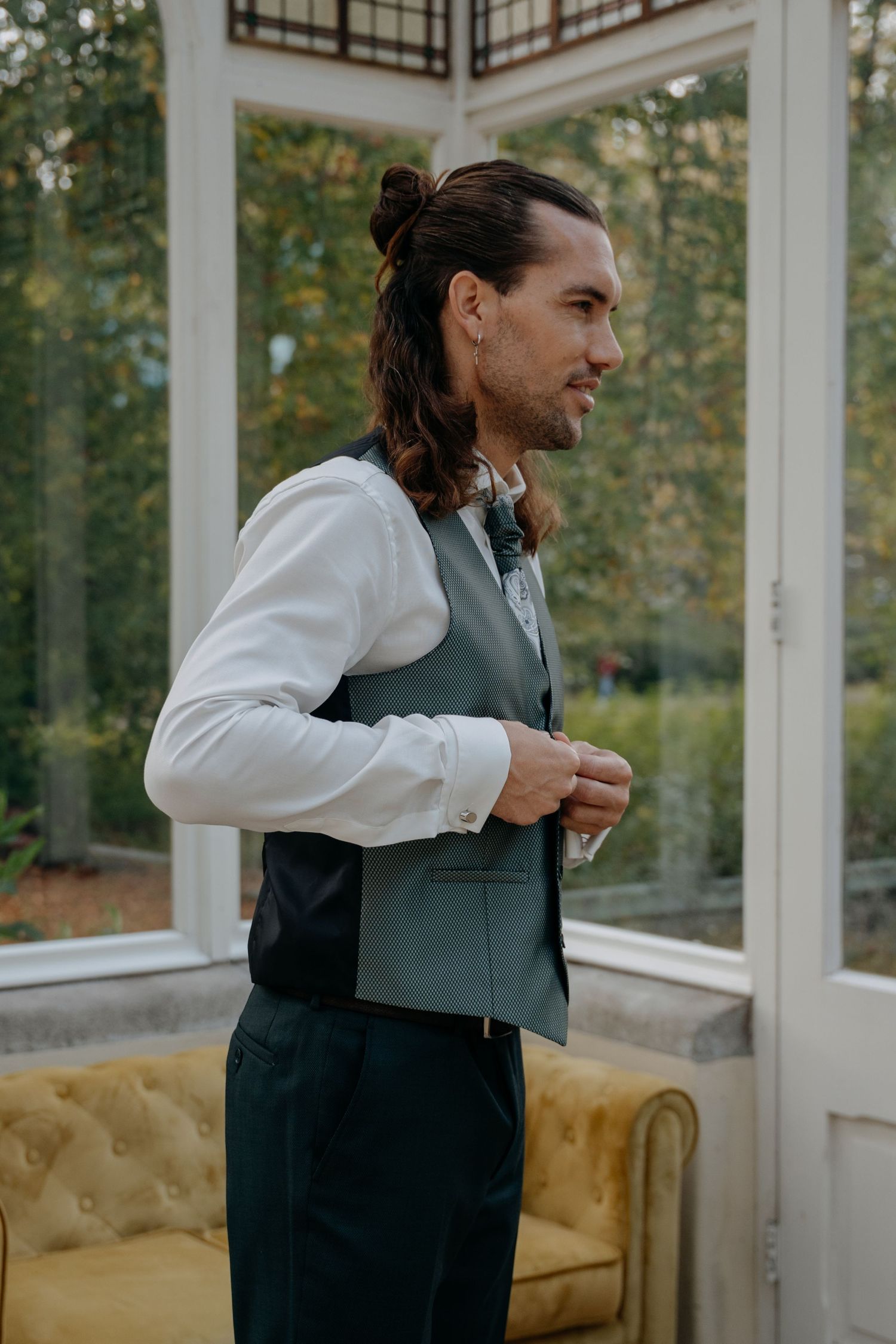 Person in formal attire moving through a sunlit glass conservatory with vintage furniture.