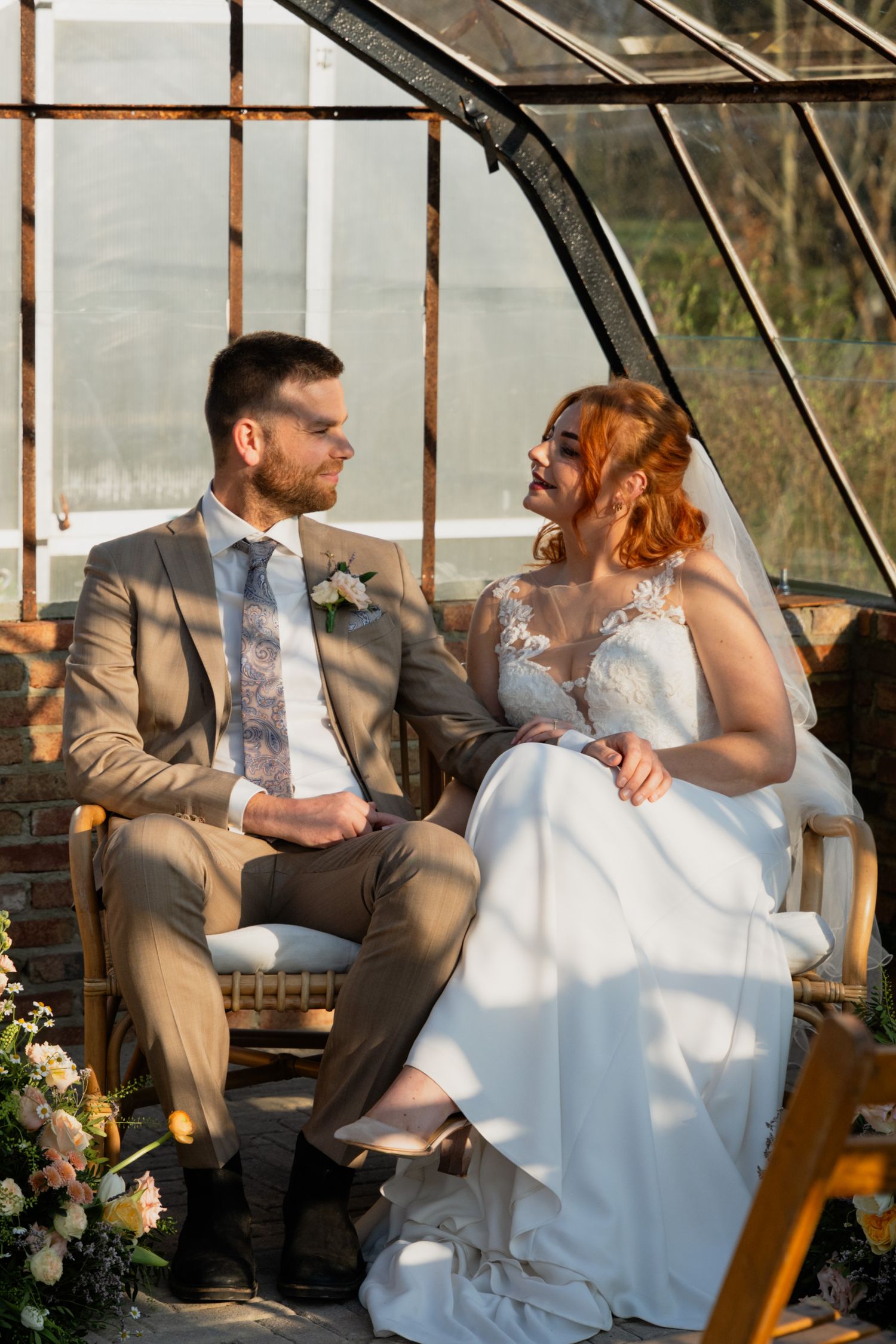 A bride and groom share a romantic moment seated together in a sunlit greenhouse, dressed in wedding attire.