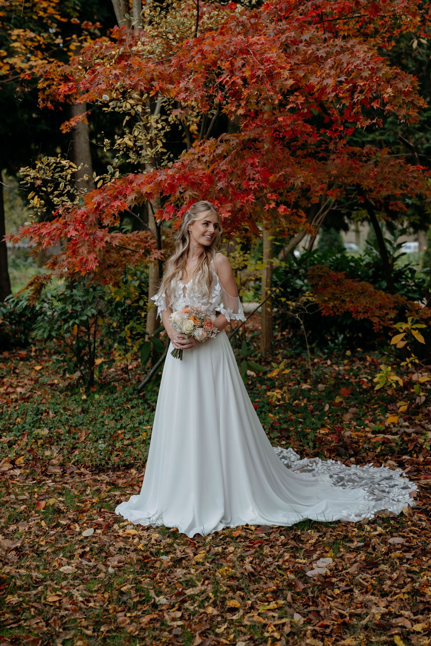 A bride in a flowing white gown poses beneath vibrant red autumn maple trees.