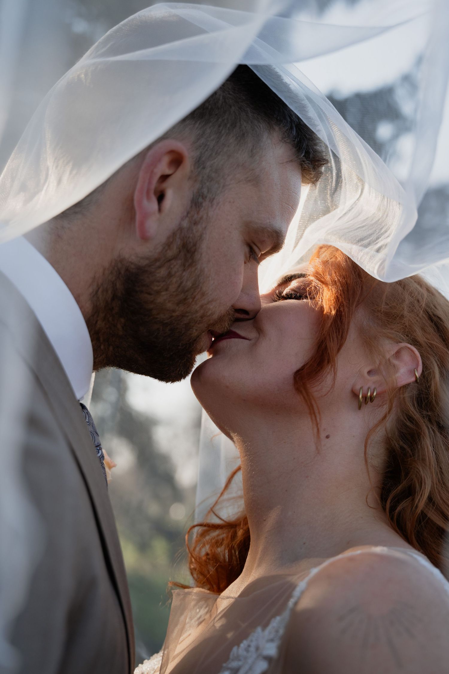 A bride and groom share a tender kiss under a flowing white veil, bathed in warm golden sunlight.