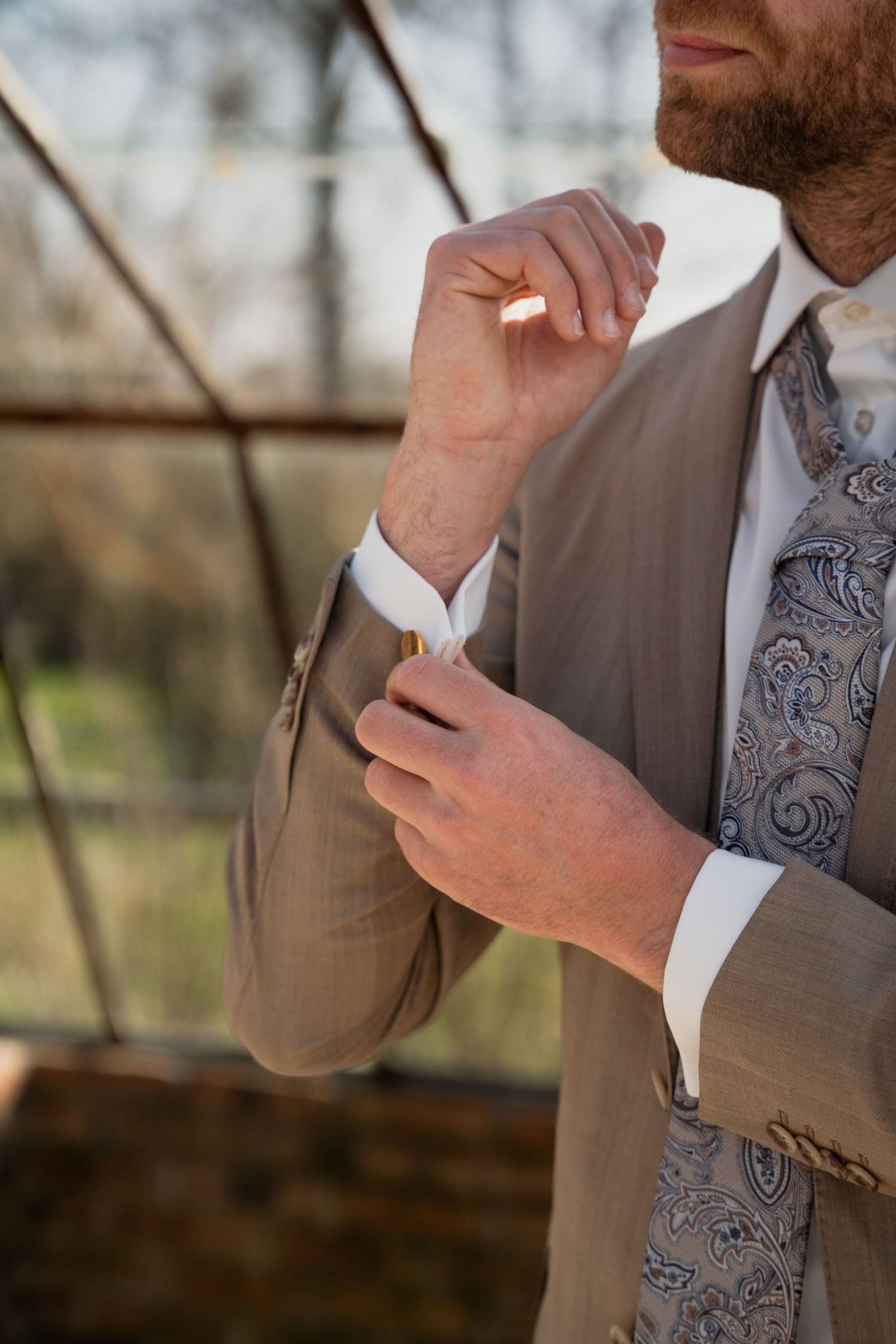 Bearded groom in tan suit and paisley tie fastening cufflinks outdoors.