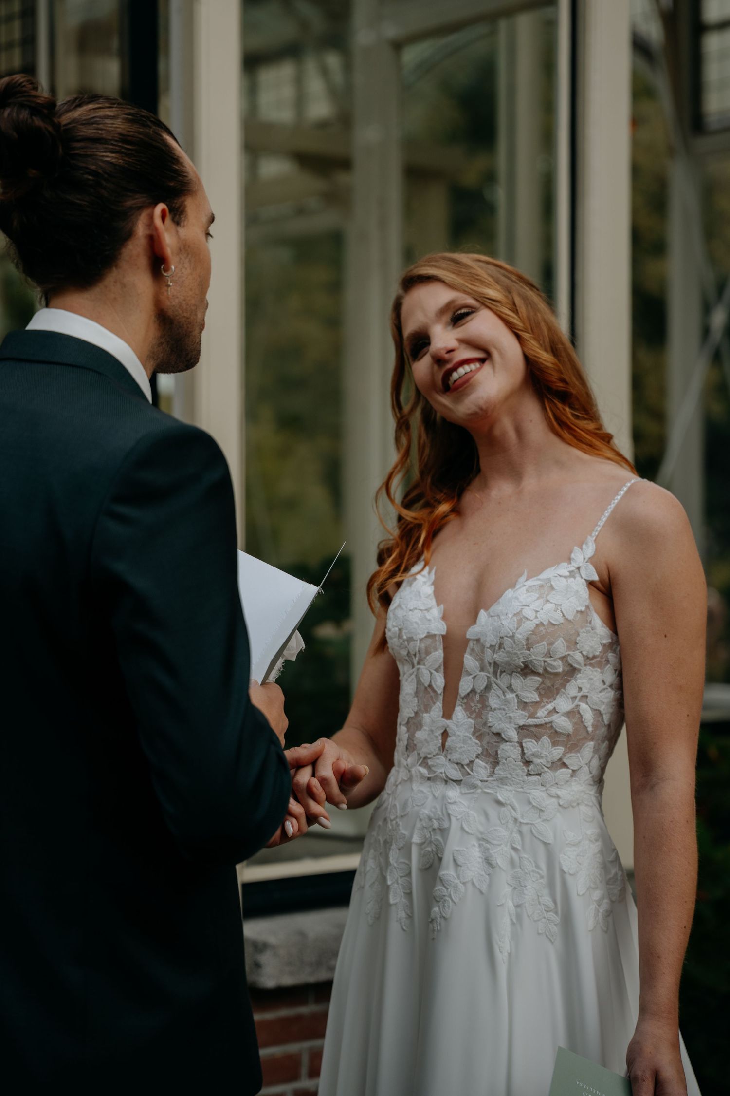 Couple sharing a candid moment in their wedding attire by glass windows.