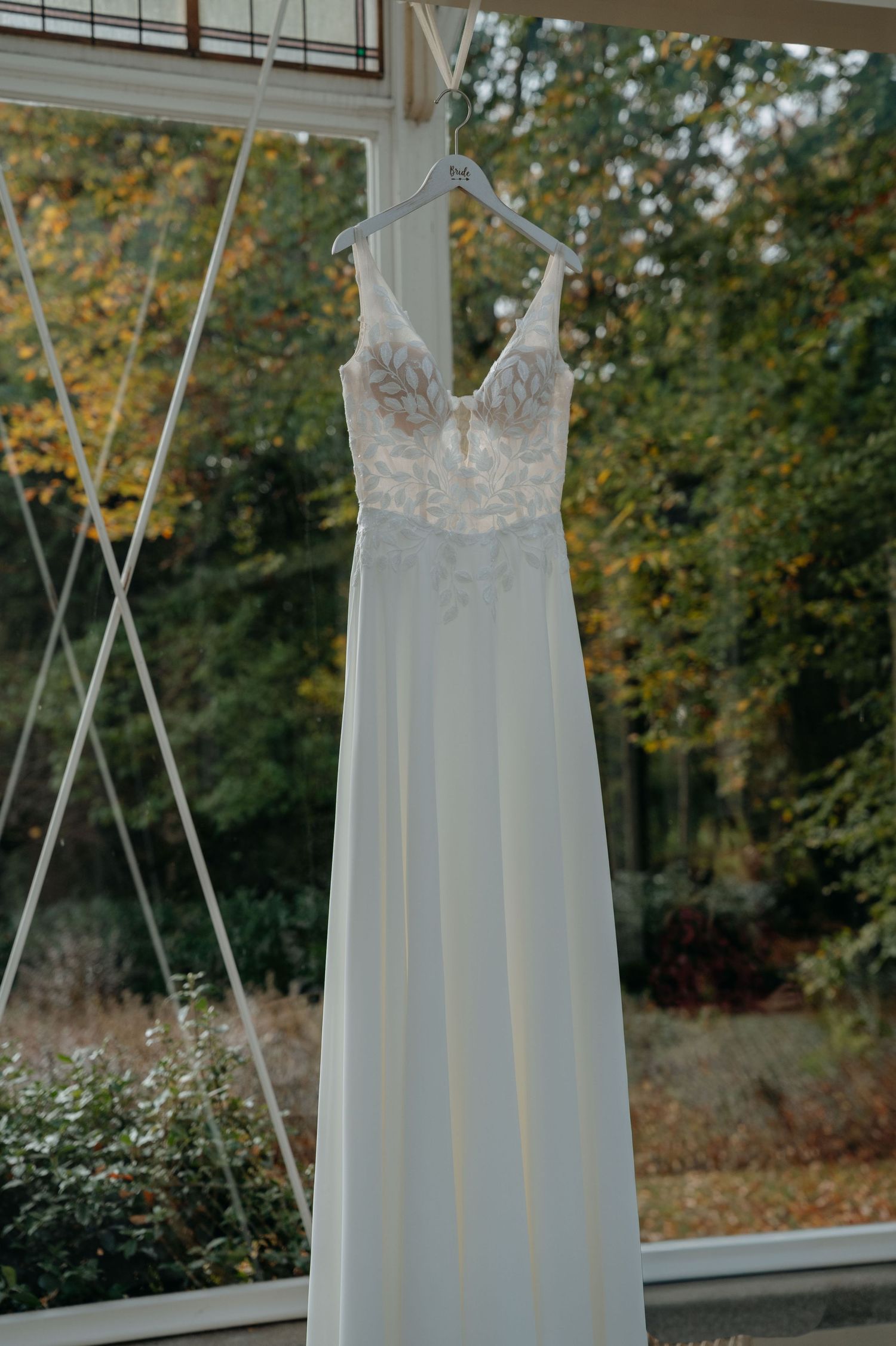 An elegant white wedding dress with lace bodice and flowing chiffon skirt hangs by straps against a window with trees outside.