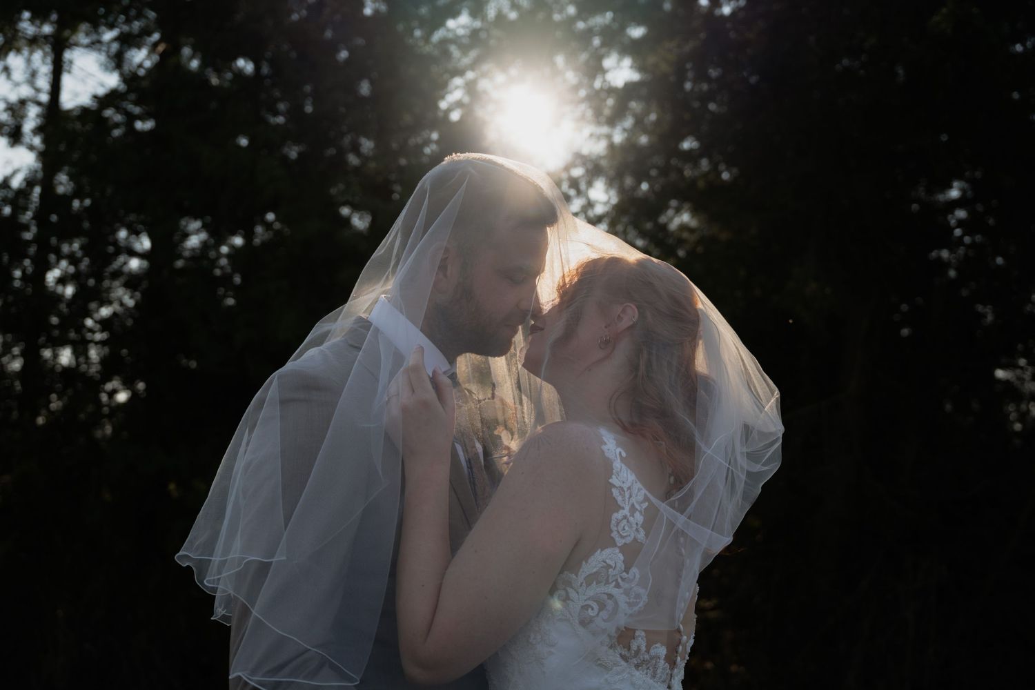 A bride and groom share a romantic moment under a veil with sunlight glowing behind them outdoors.