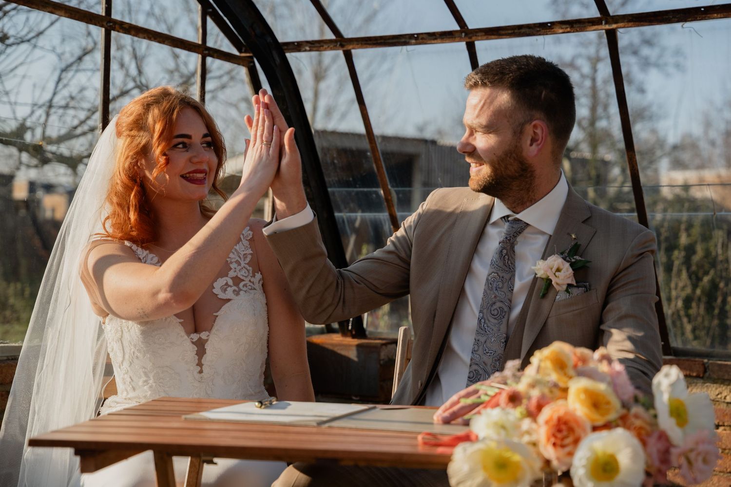Joyful newlyweds high-fiving at wedding signing table in a sunlit glass greenhouse venue.