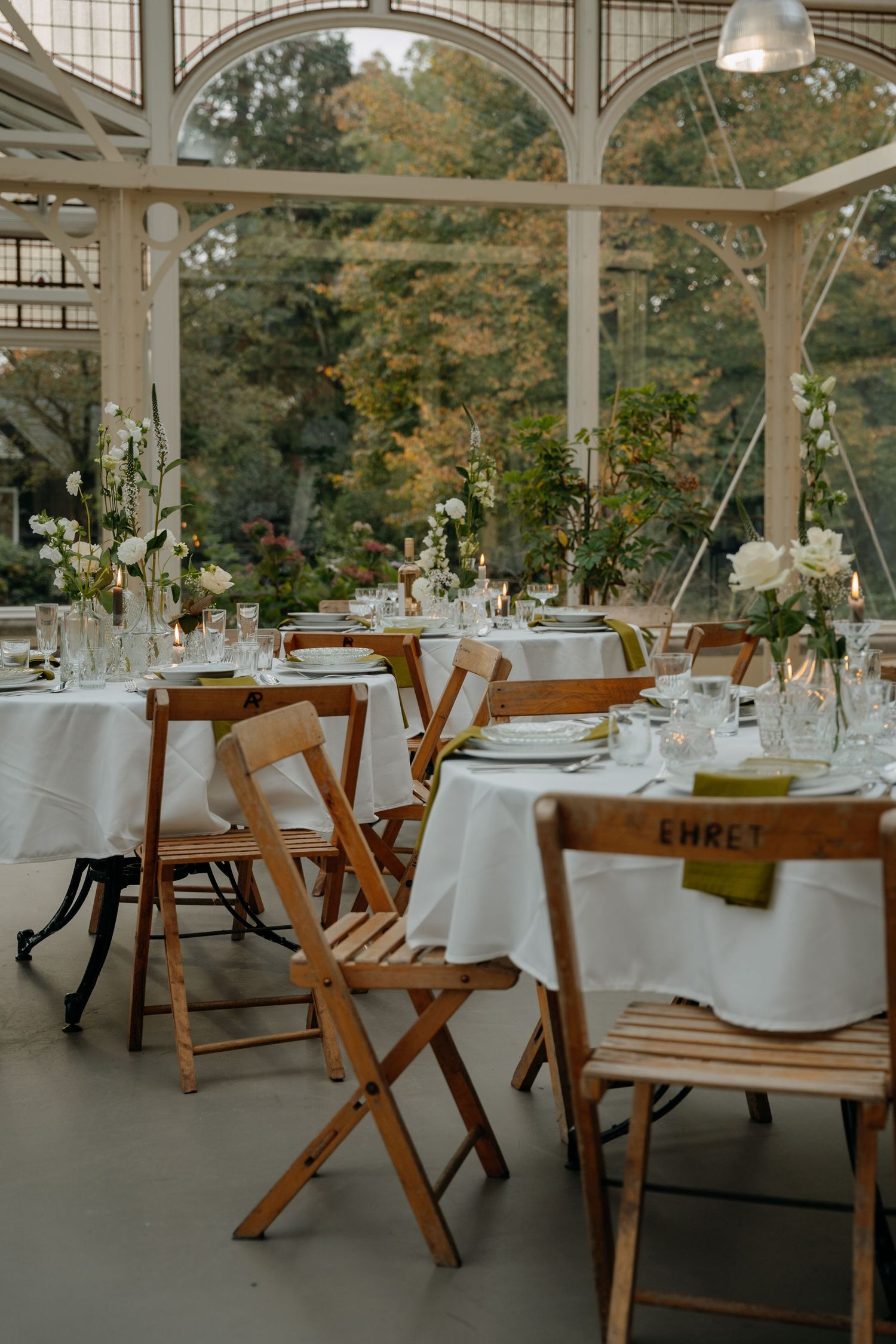 An elegant wedding reception venue with white tablecloths and wooden chairs in a glass-enclosed conservatory.