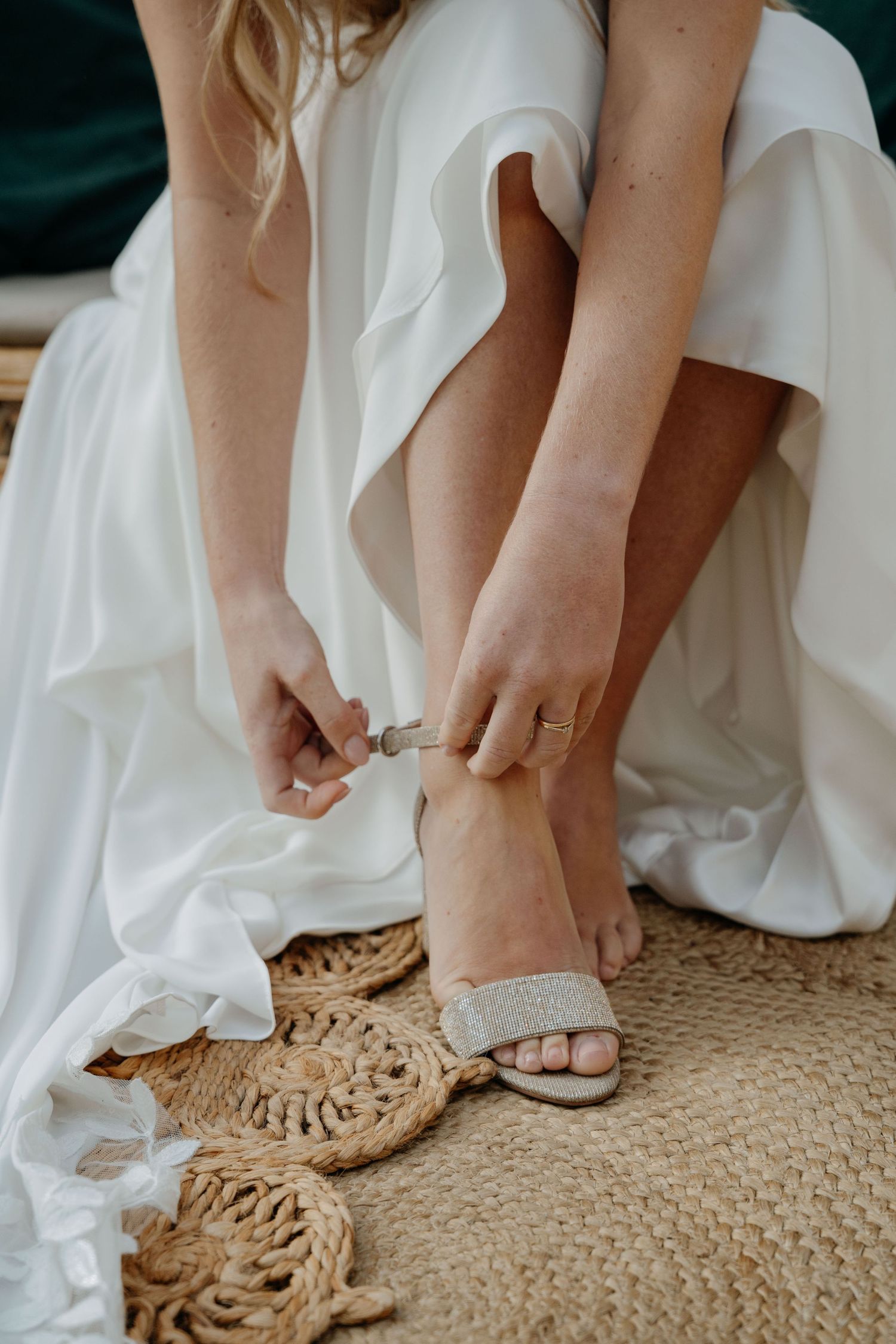 Bride wearing delicate white wedding flats puts on her shoes while sitting on a textured jute rug.