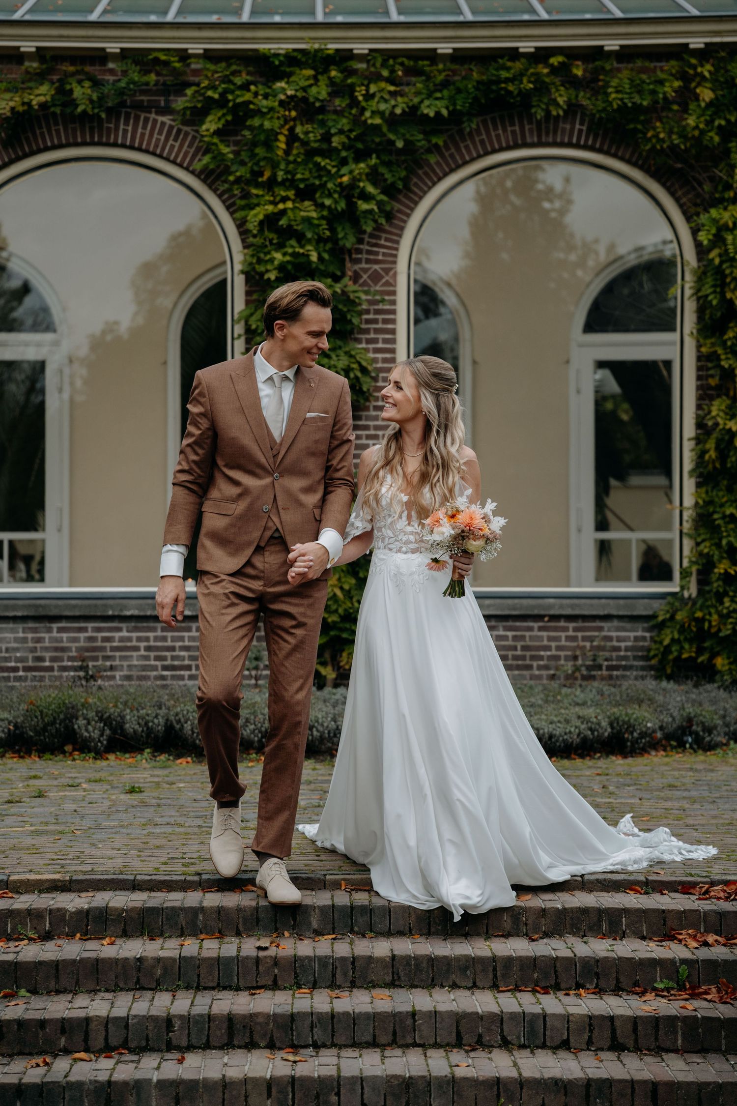 A couple in wedding attire stands on brick steps outside a historic building with ivy-covered arches.