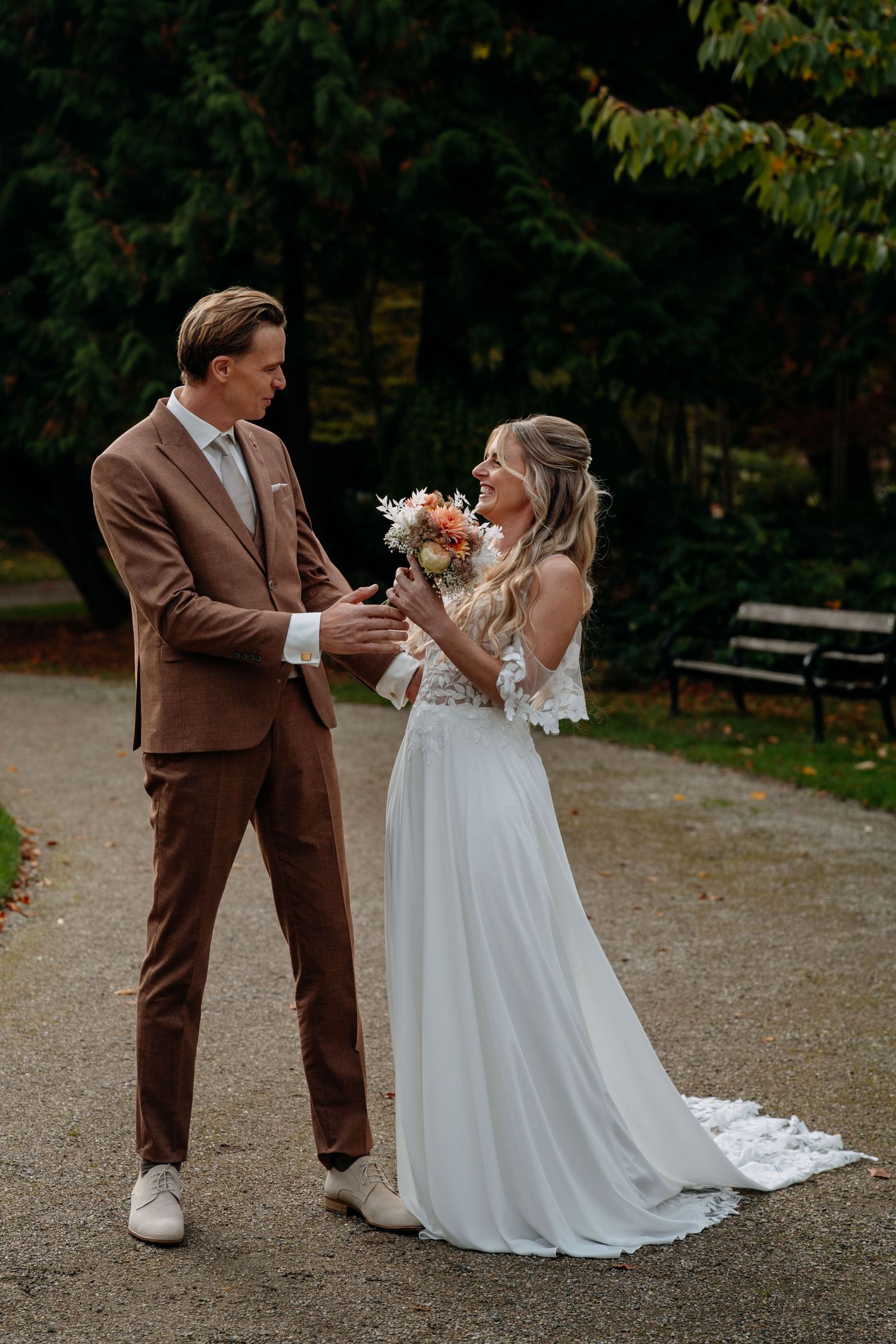 Bride and groom share playful moments on a gravel path surrounded by autumn trees.