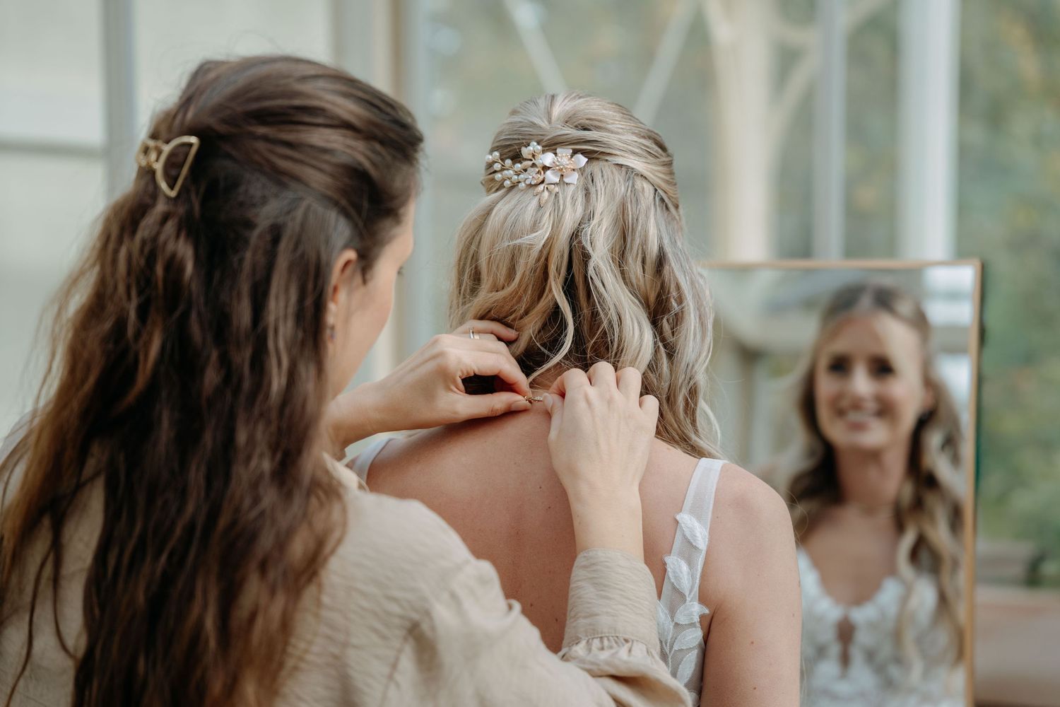 A stylist arranges a bride's hair with delicate floral accessories in a sunlit room while another person looks on.
